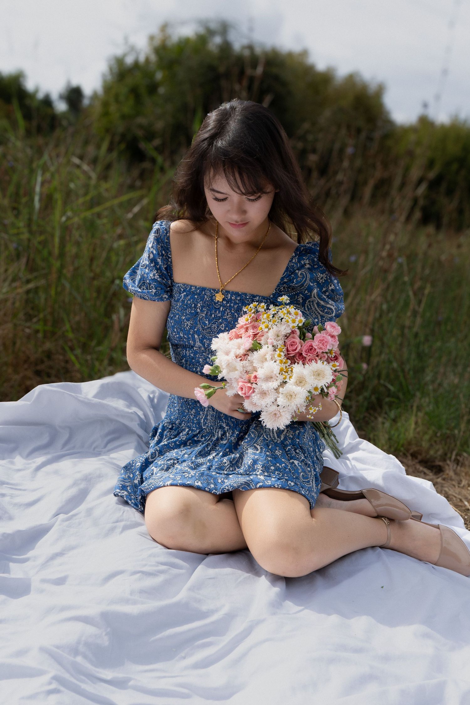 Young woman in blue floral dress sitting on white blanket outdoors, holding a bouquet of pink and white flowers.