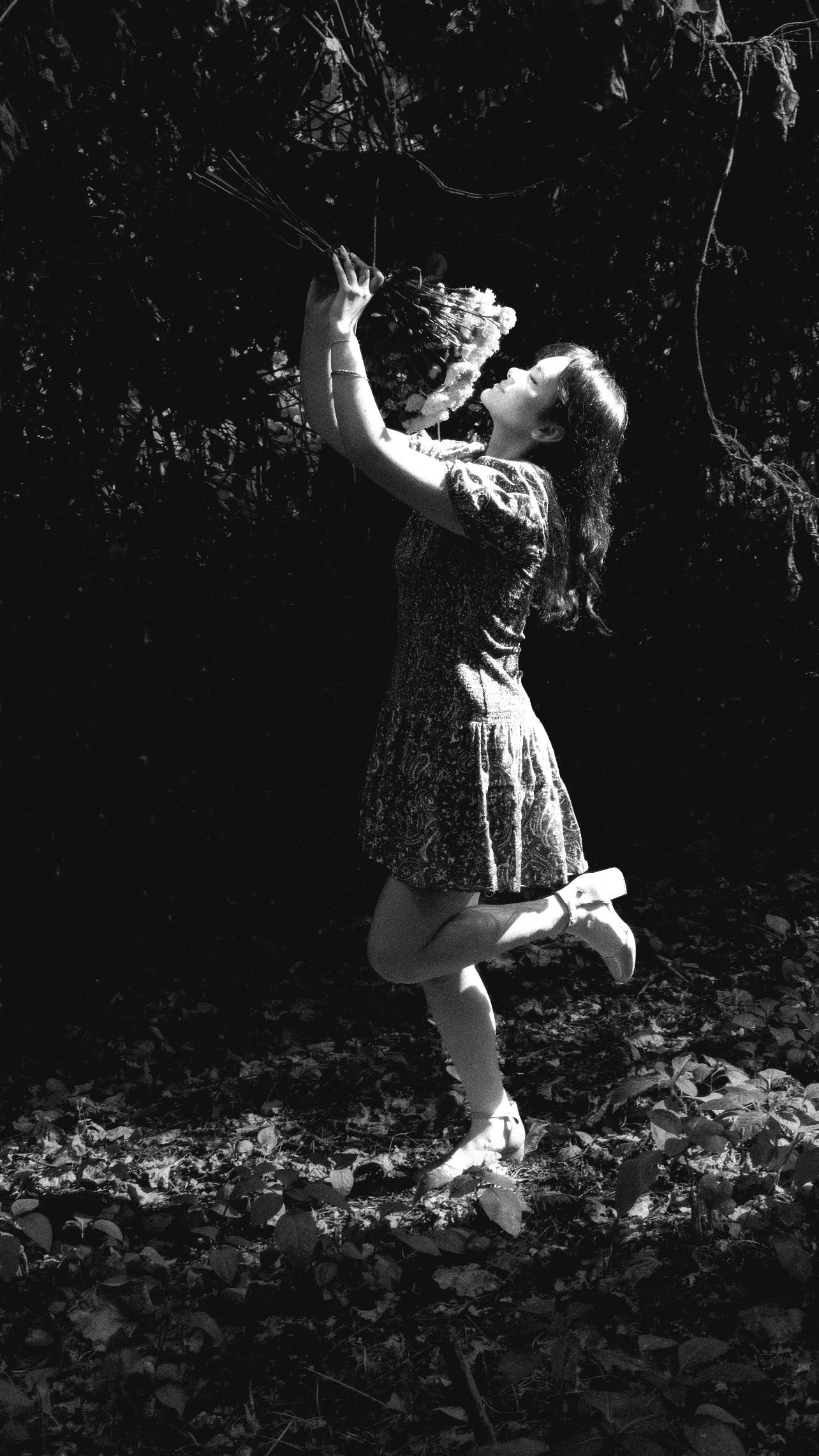 Black and white photo of a girl in a dress reaching up with flowers in a dark forest setting.