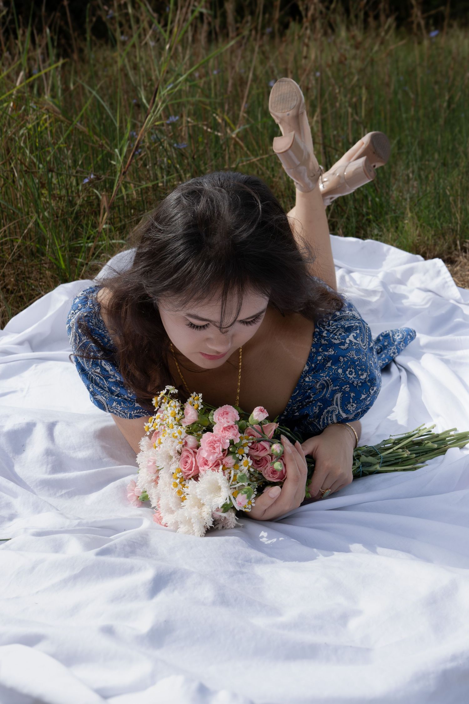 Young woman in blue floral dress lying on white blanket outdoors, holding pink and white flower bouquet.