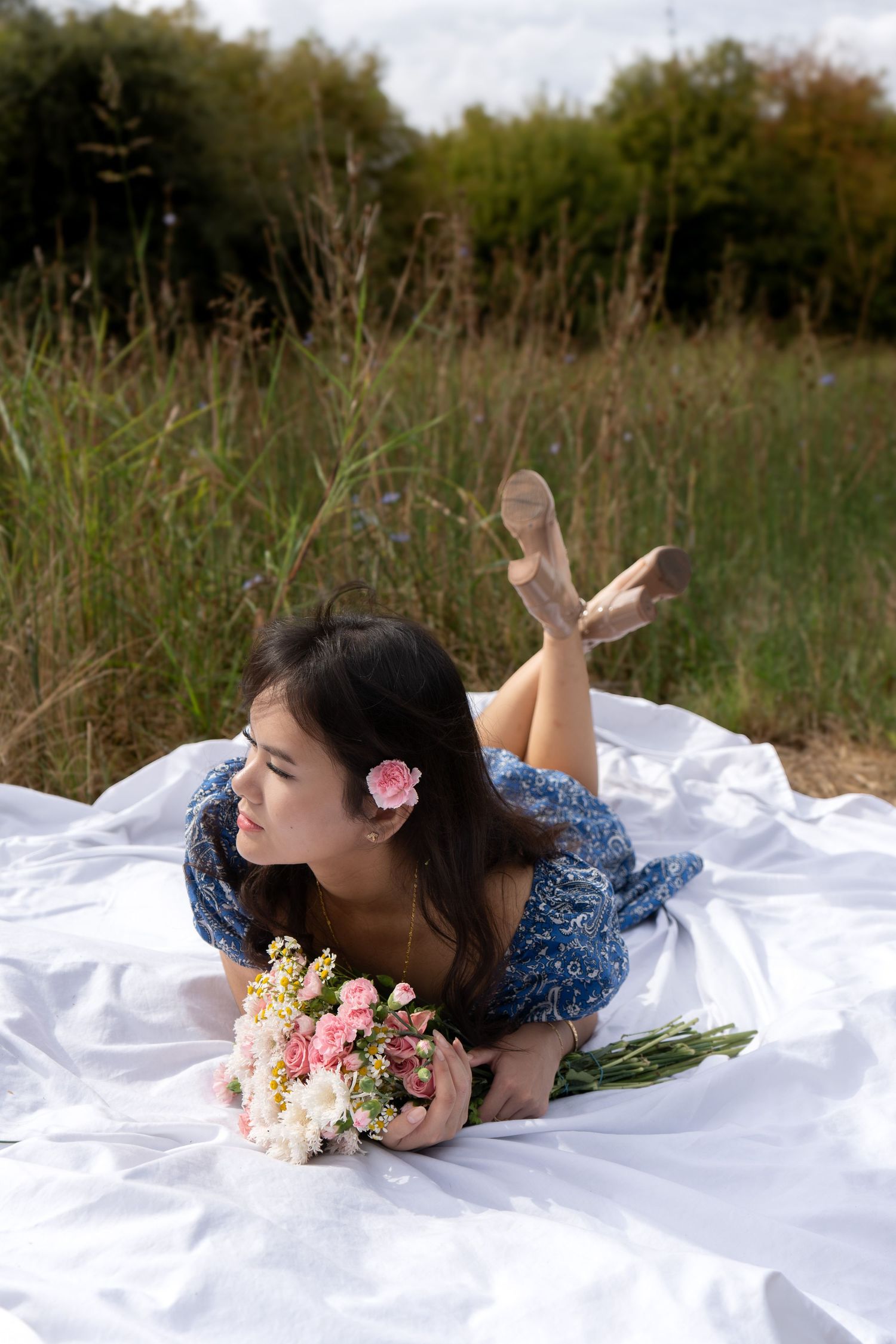 Brunette woman in blue dress lying on white blanket in field, holding bouquet with pink flower in hair.