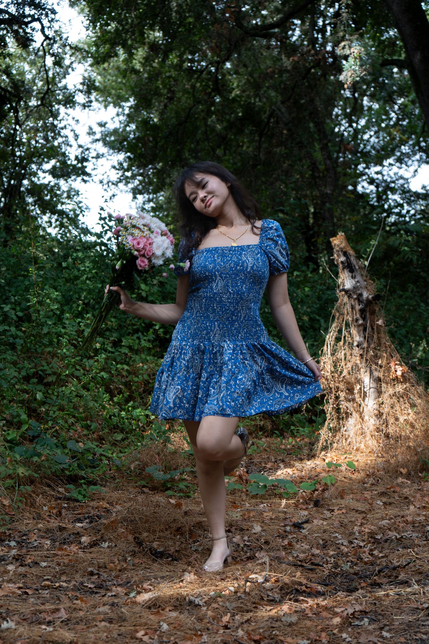 Young woman in blue floral dress holding pink bouquet, posing barefoot in a lush green forest setting.