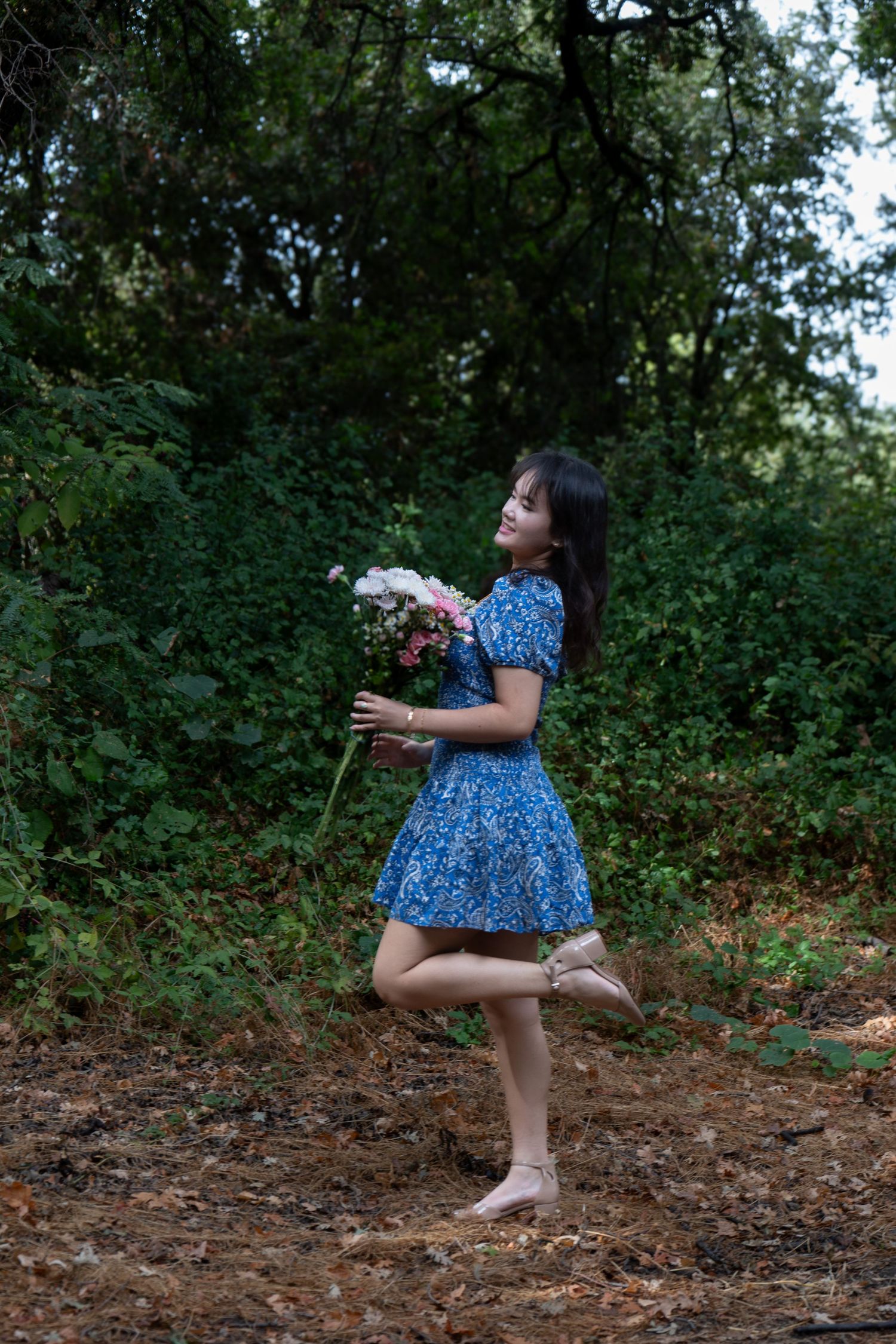 Young woman in blue floral dress playfully posing with bouquet of flowers in lush green forest.