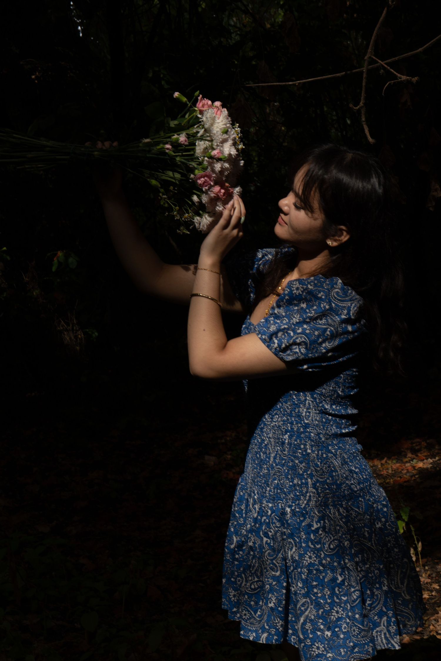 Young woman in blue floral dress holding pink wildflowers bouquet in dappled sunlight against dark forest background.