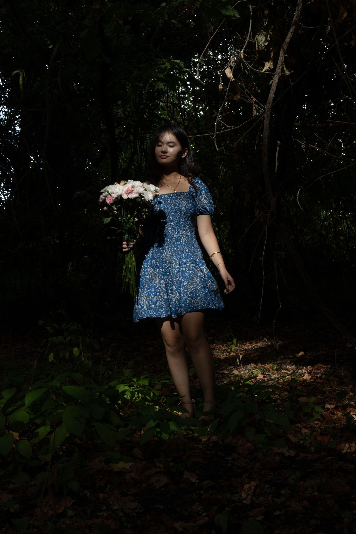 Young woman in blue floral dress holding bouquet of pink and white flowers in dark forest setting.