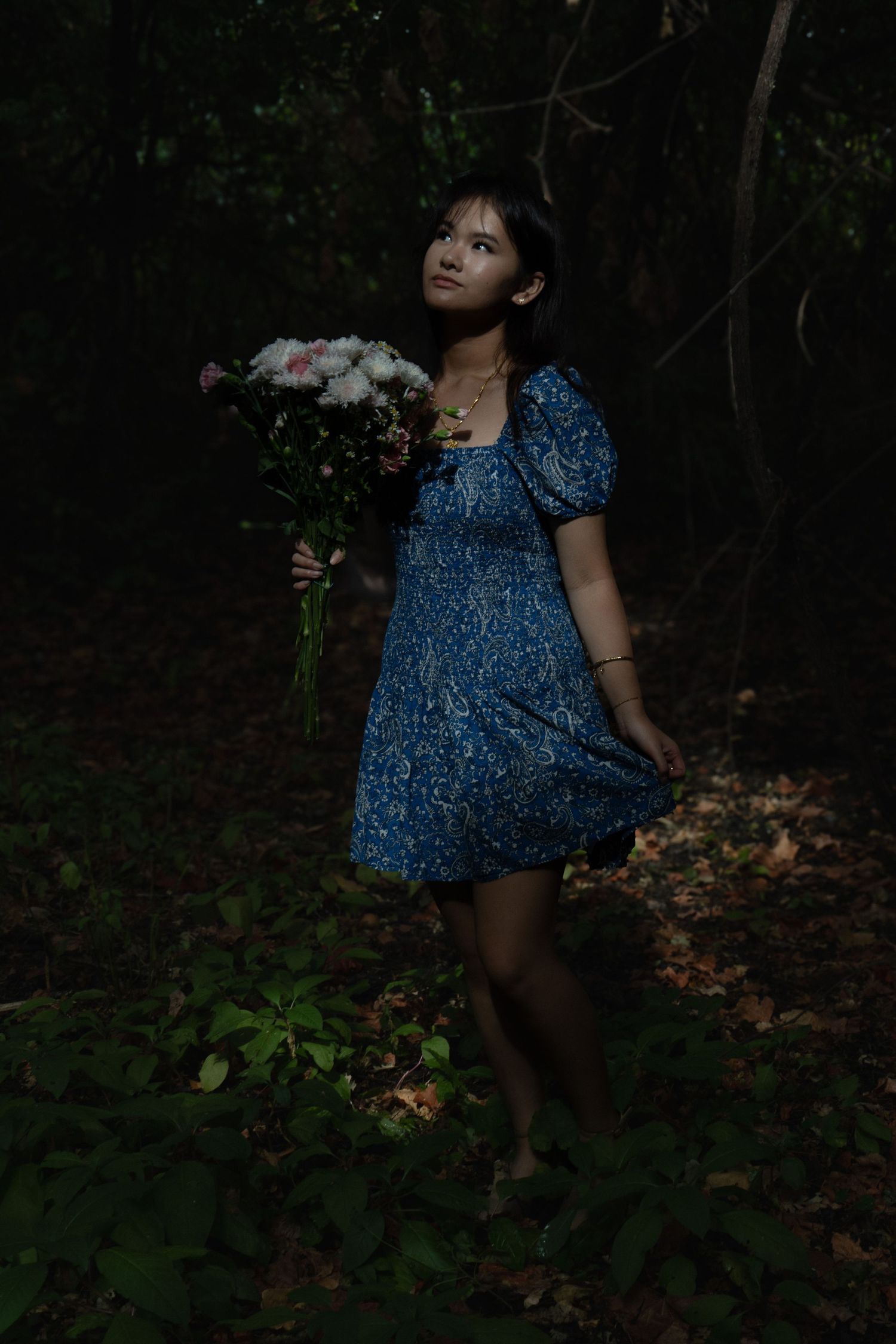 Young woman in blue patterned dress holding flower bouquet while standing in dark forest at night.