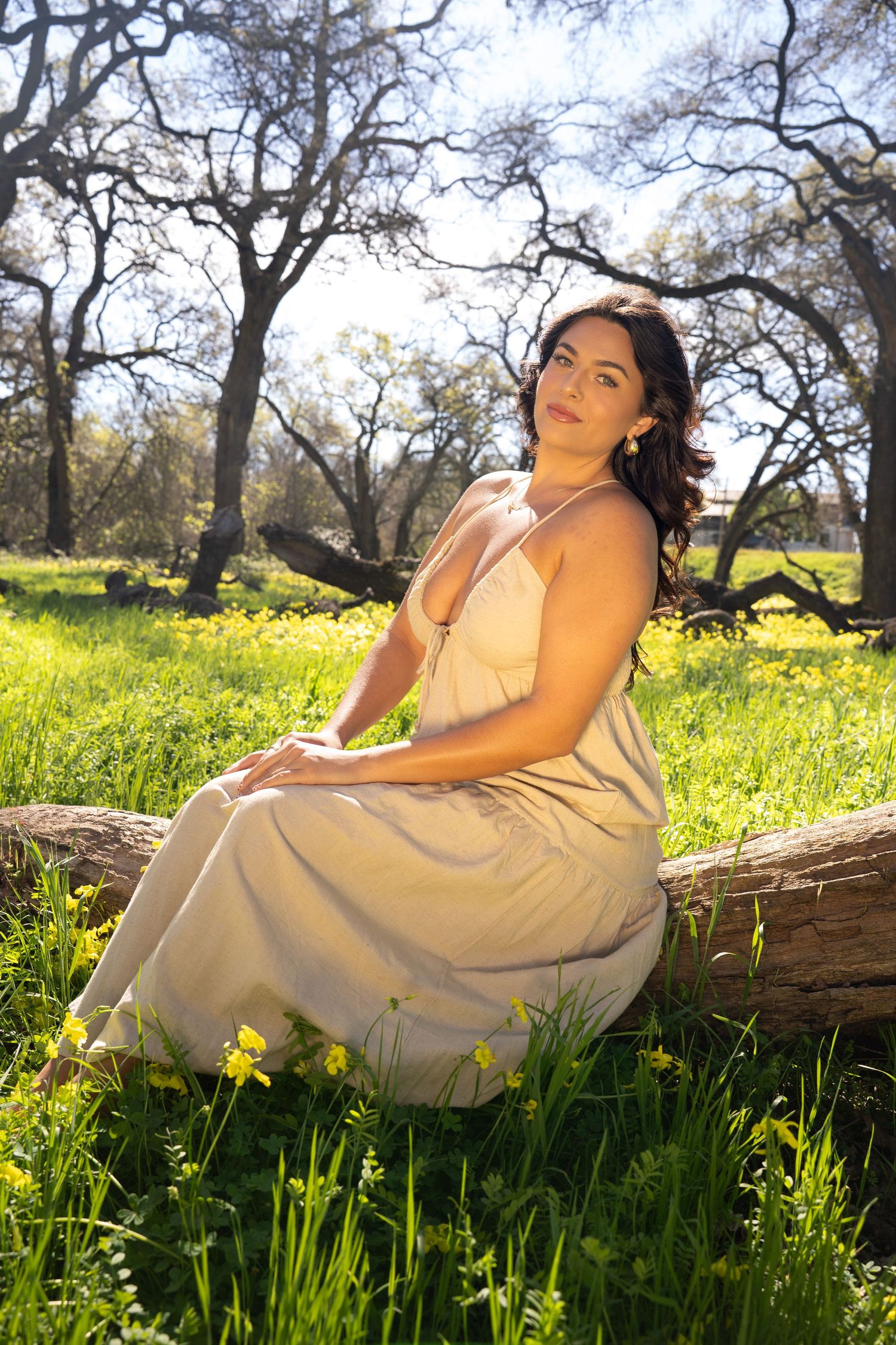 Woman in beige sundress sitting on log in sunny meadow with wildflowers and oak trees in background.