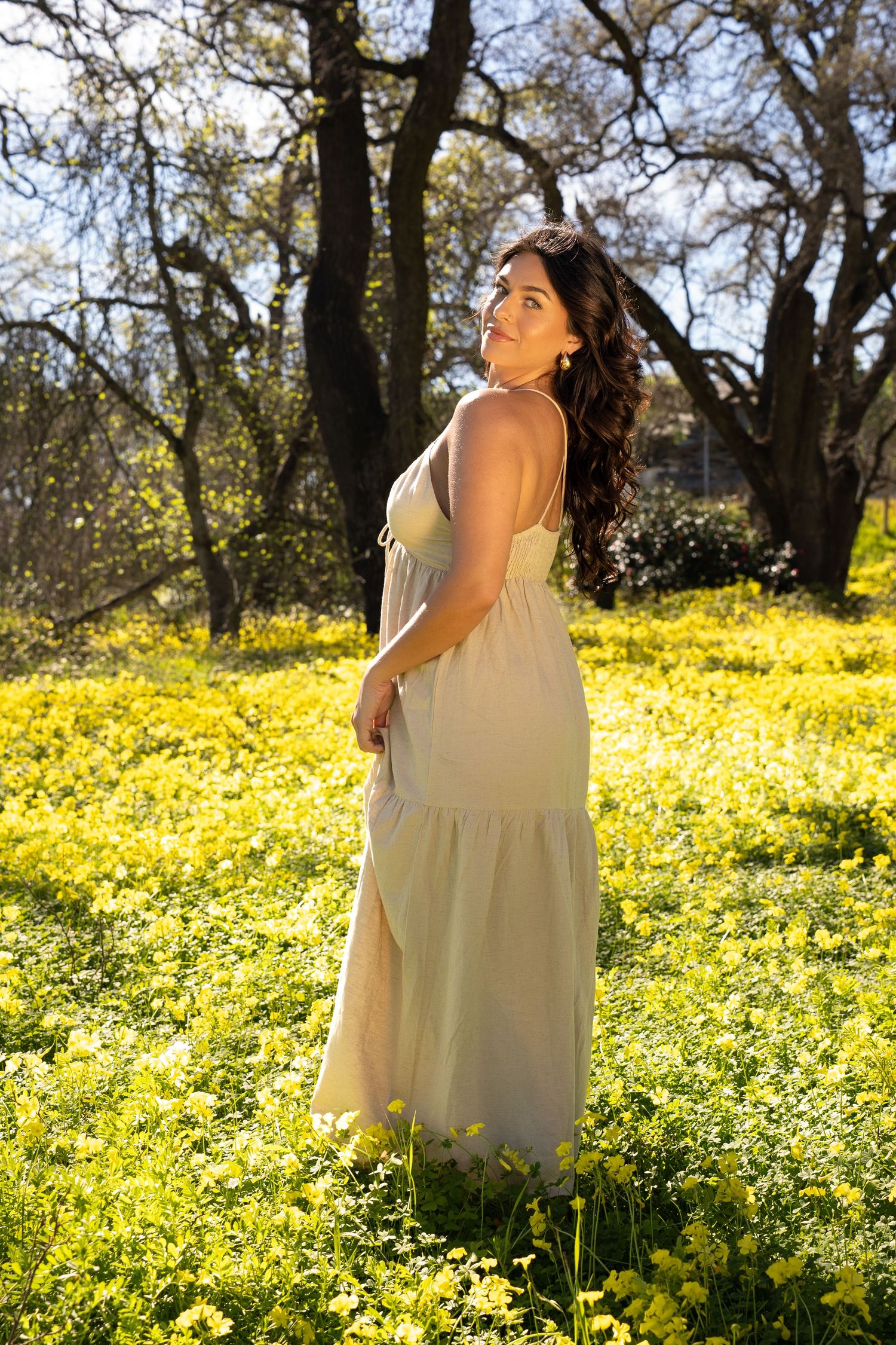 Woman in flowing yellow maxi dress standing in a sunlit field of yellow wildflowers with trees in background.