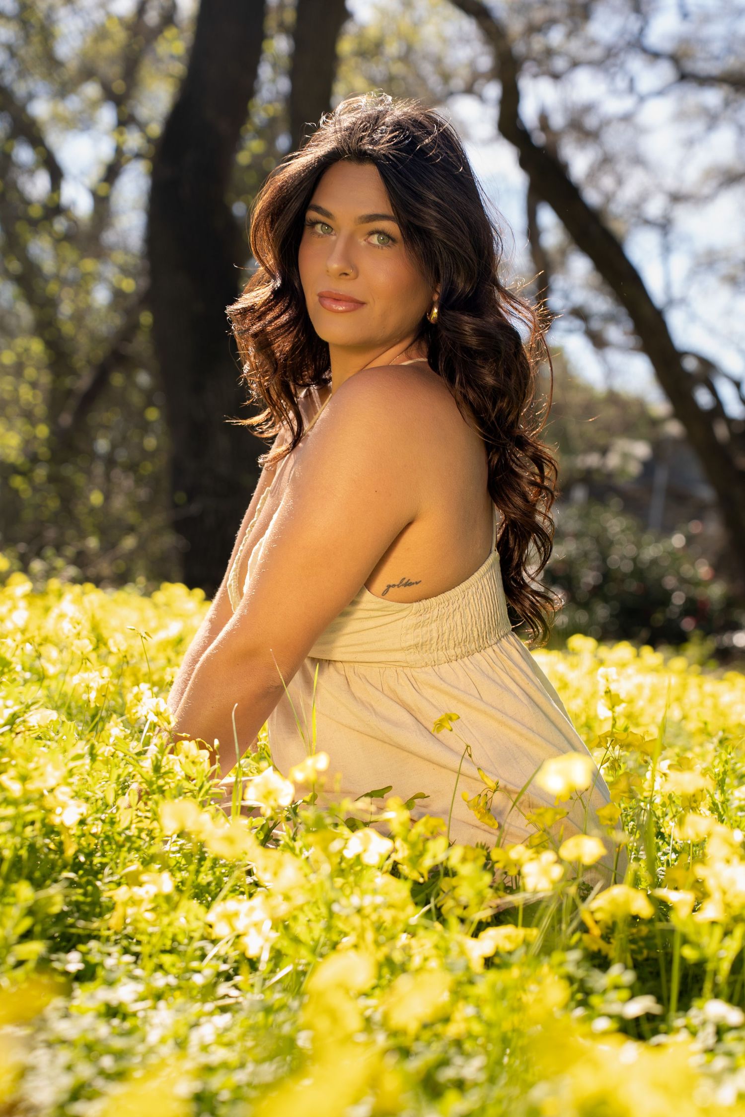 Woman in white dress sitting in yellow wildflower field, long dark curly hair, looking over shoulder, sunny outdoor portrait.