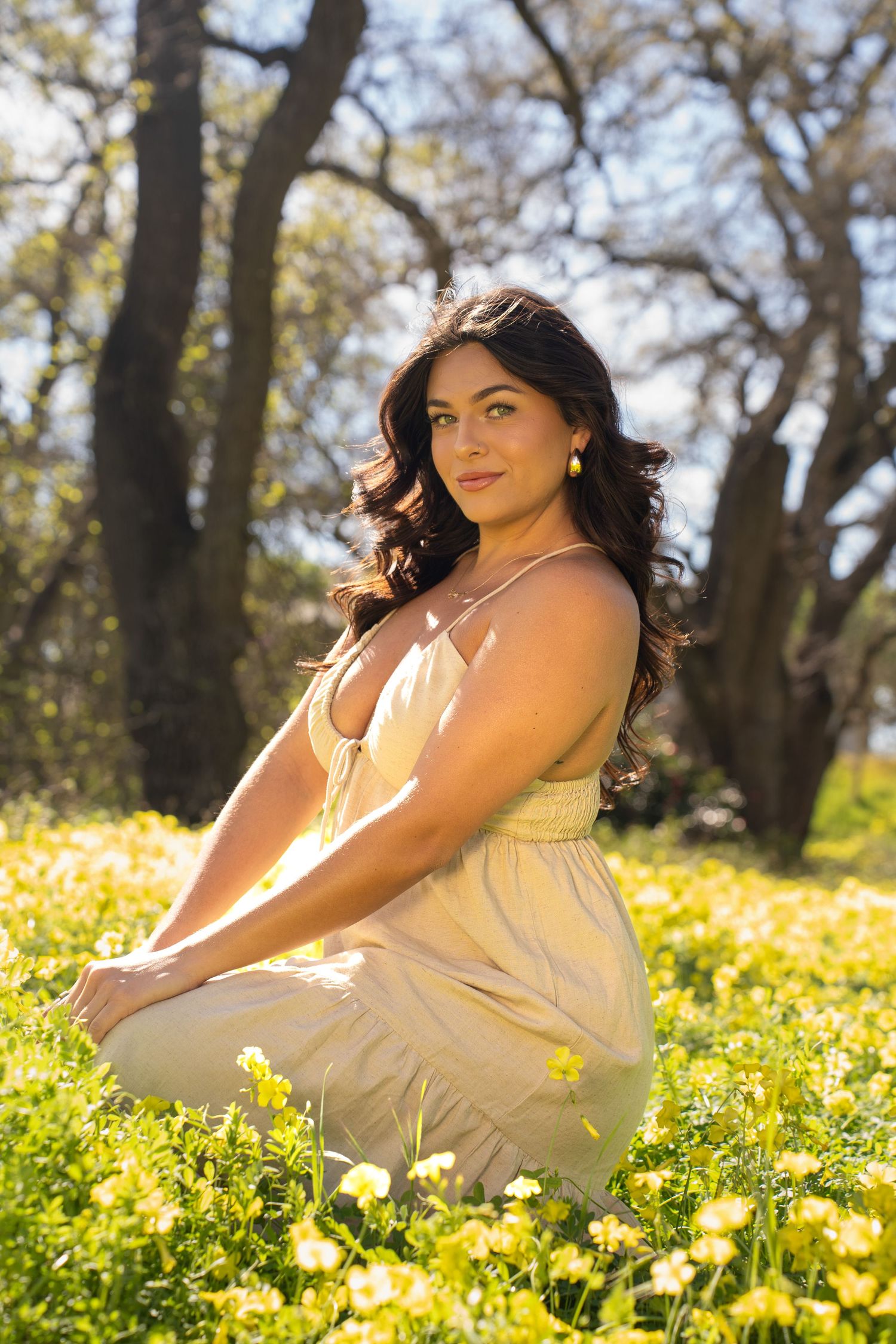 Young woman in a beige sundress sitting in a field of yellow wildflowers with trees in the background.