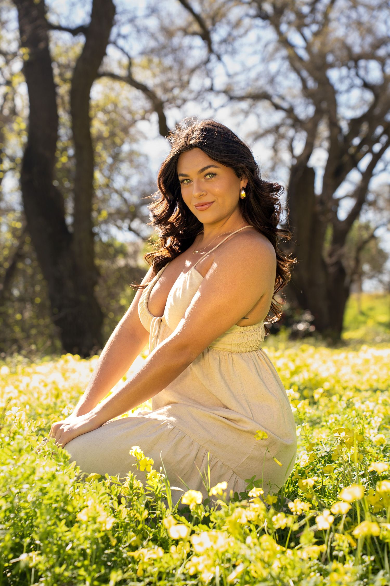 A young woman in a beige sundress sits gracefully among yellow wildflowers in a sunlit meadow with trees behind her.