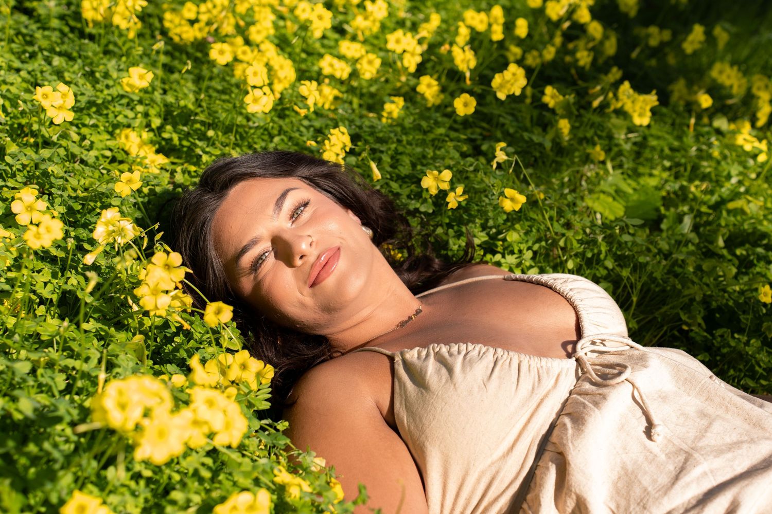 A relaxed woman lying among vibrant yellow wildflowers in a sunny meadow, wearing a light dress with eyes closed.