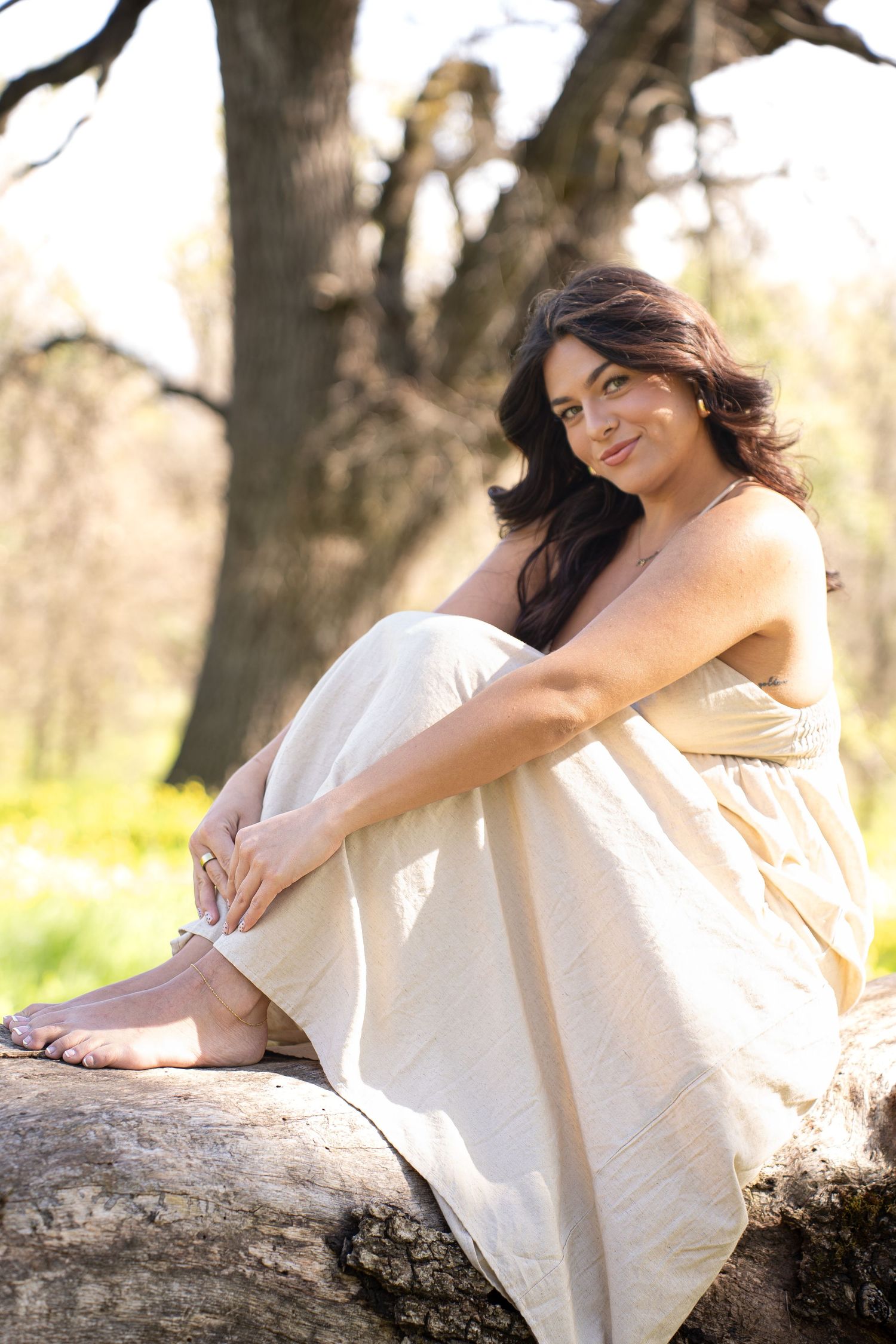 Barefoot woman in cream dress sitting on log in sunlit forest, smiling naturally at camera.