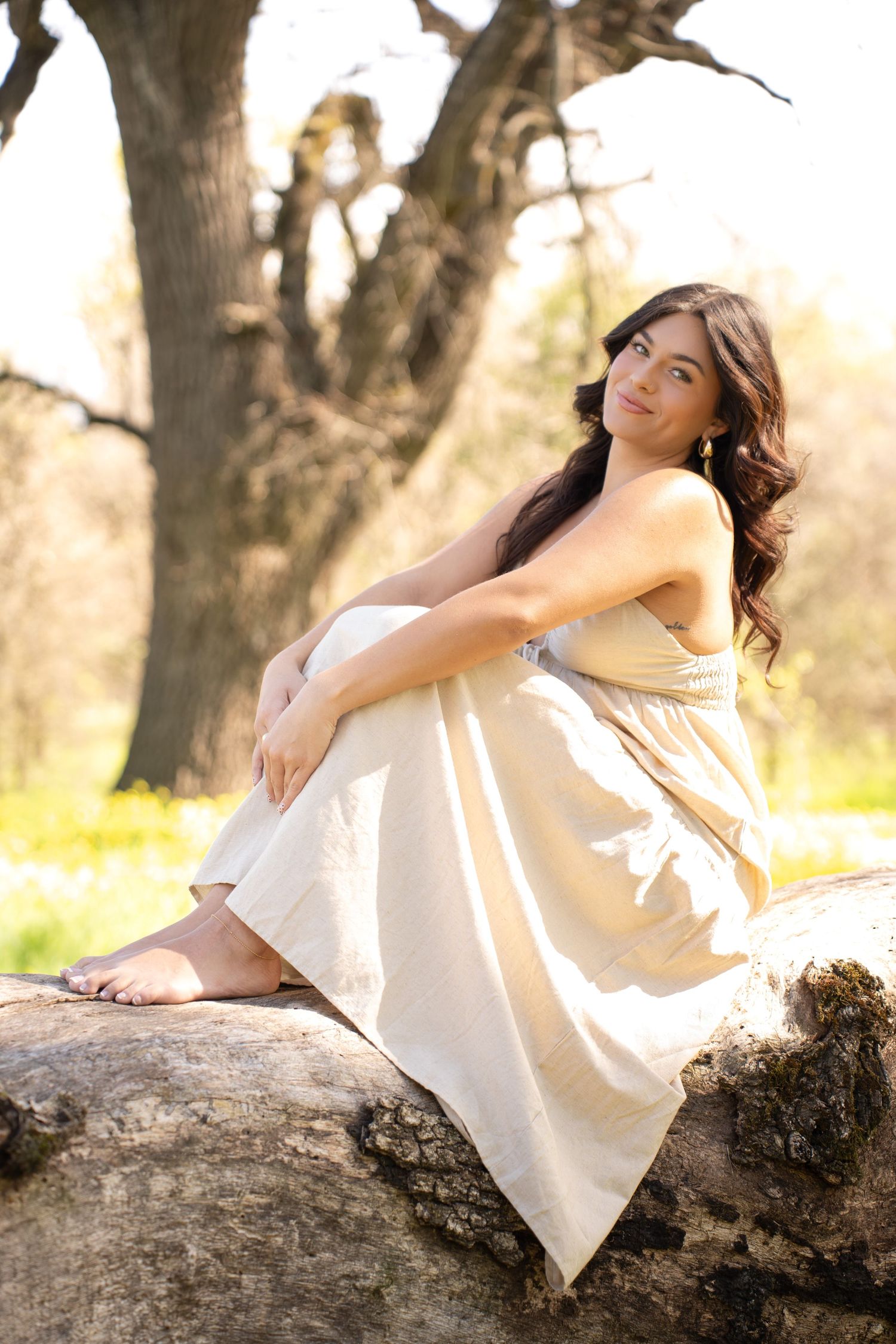 Young woman in cream dress sitting barefoot on a rock outdoors, surrounded by trees and wildflowers in soft sunlight.