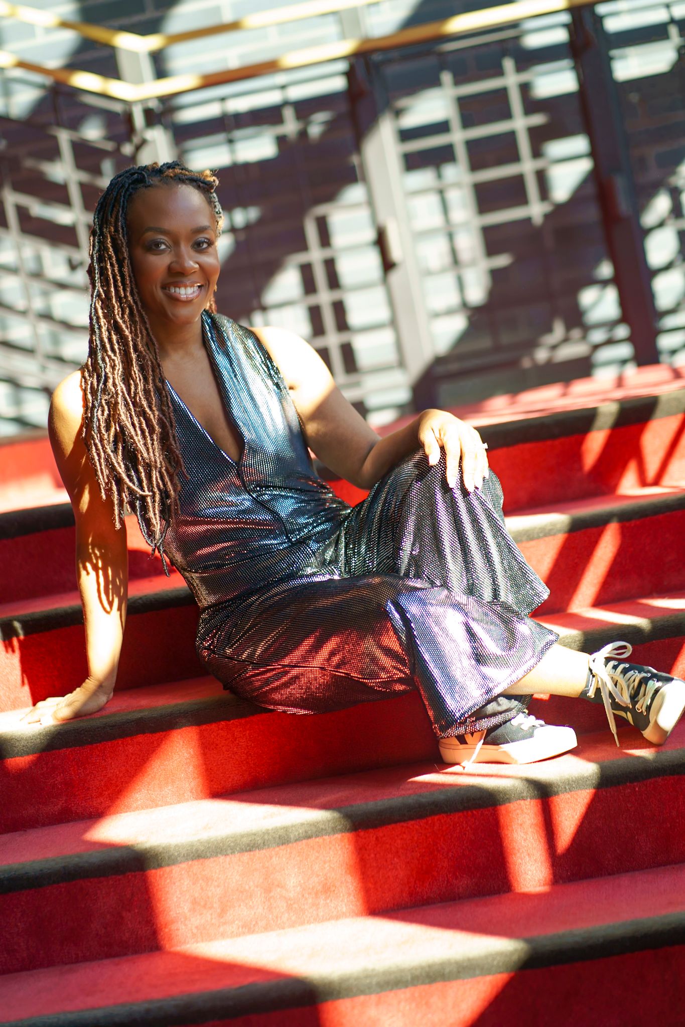 Smiling woman with braids wearing a shiny holographic jumpsuit sitting on red stadium steps in sunlight.