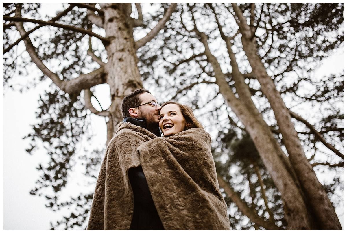 Couple wrapped in brown blanket laughing together outdoors among tall trees in a cozy winter engagement photo.