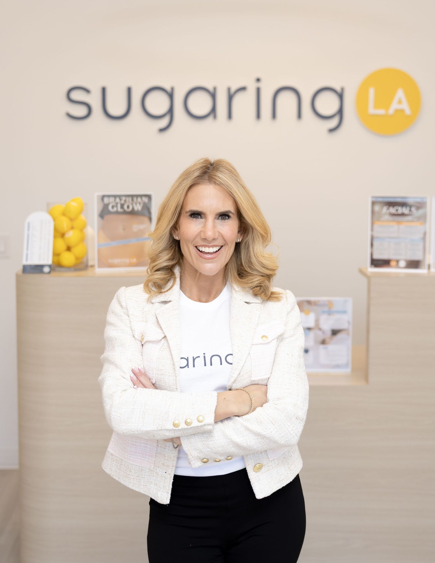 Professional in a white blazer and black skirt stands confidently at Sugaring LA salon with branded signage visible.