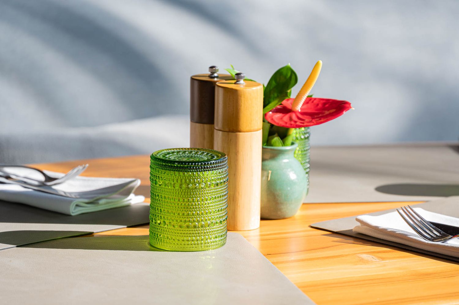 Outdoor dining table with green glass jar, wooden pepper mill, and floral centerpiece in bright sunlight.