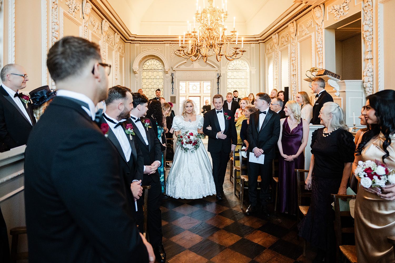 Bride walks down the aisle in elegant ballgown at a grand chandelier-lit wedding ceremony venue.