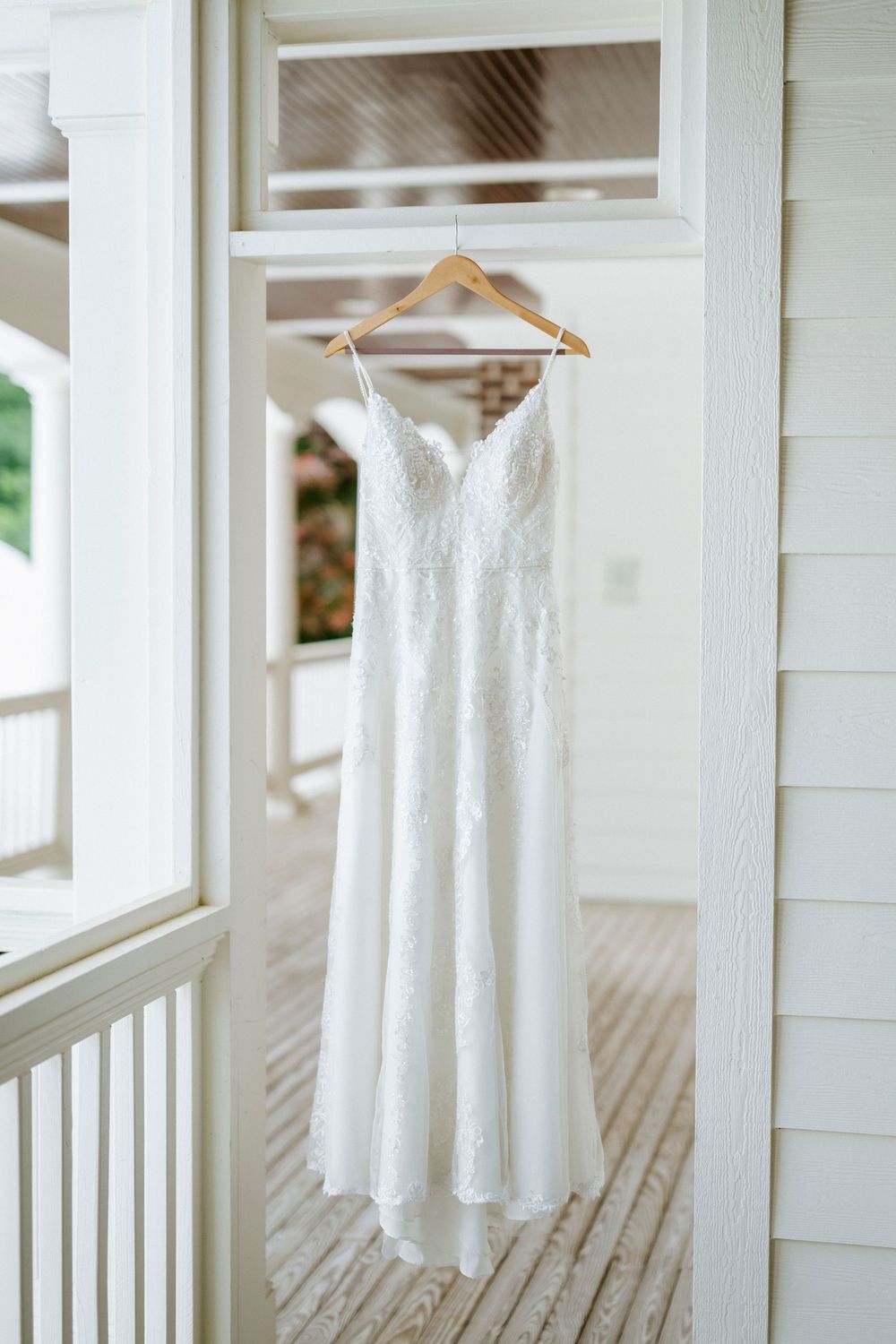 White lace wedding dress with spaghetti straps hanging on a wooden hanger in a bright white doorway.