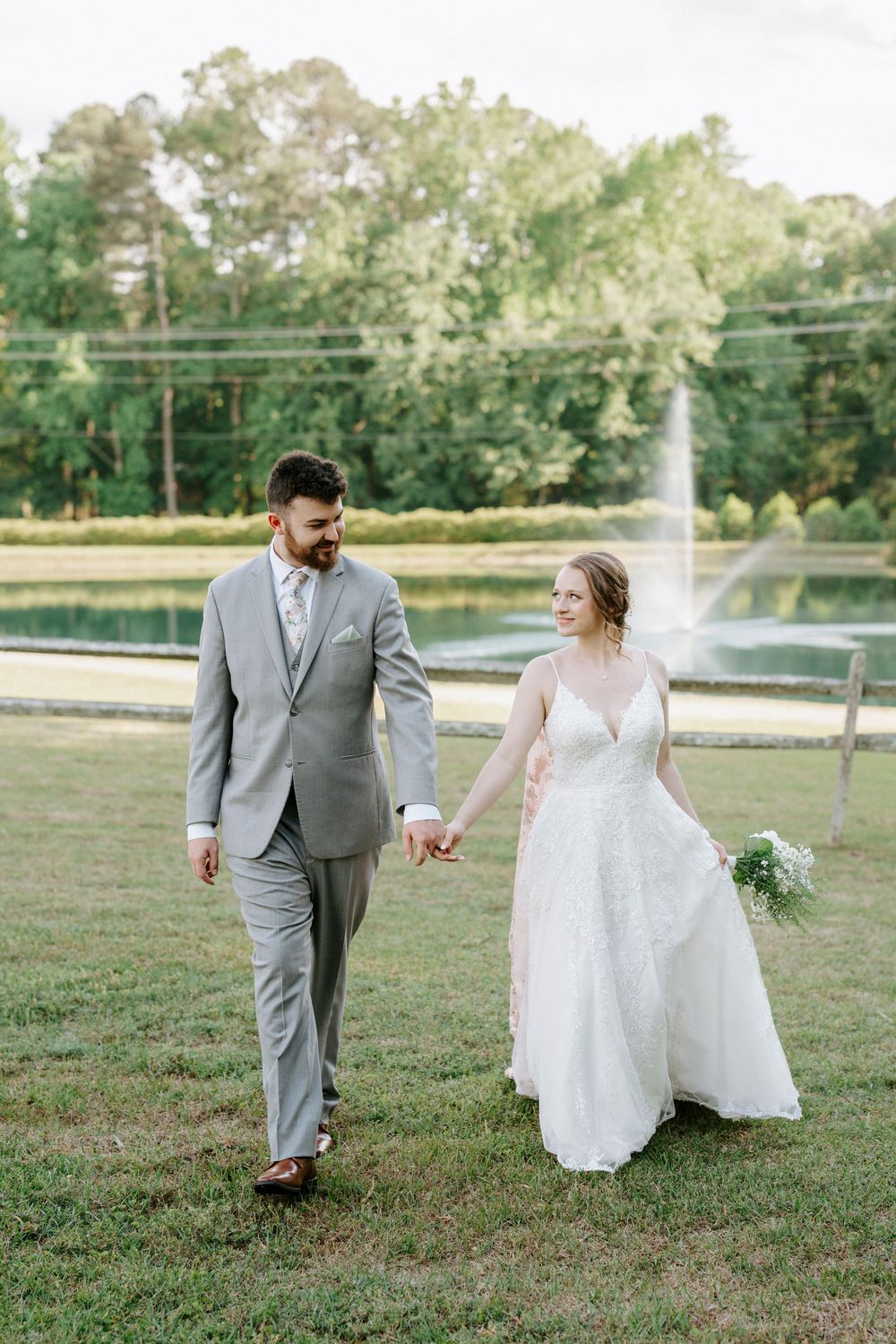 A bride and groom walk hand in hand on green grass with a pond fountain backdrop on their wedding day.