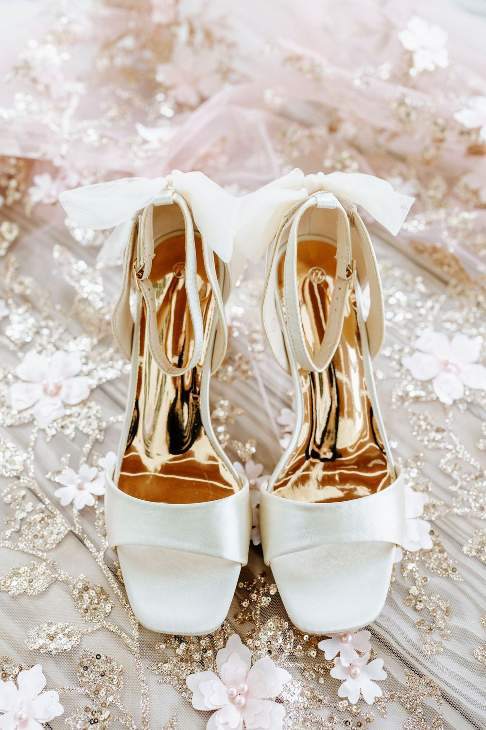 Elegant white satin bridal heels with gold insoles and ribbon ankle ties displayed on floral embroidered fabric.