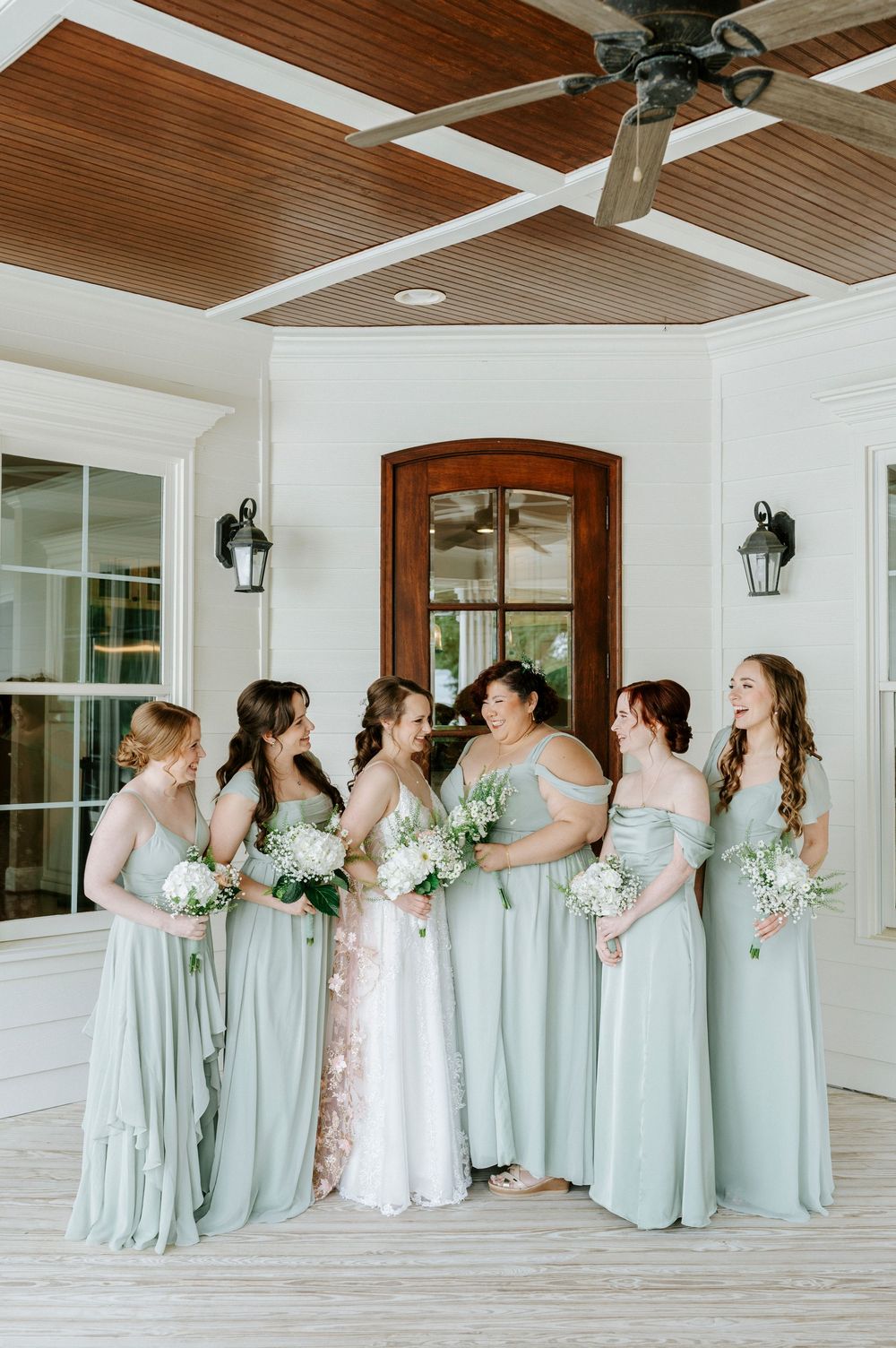 Bride surrounded by bridesmaids in sage green gowns holding white bouquets on a covered porch.