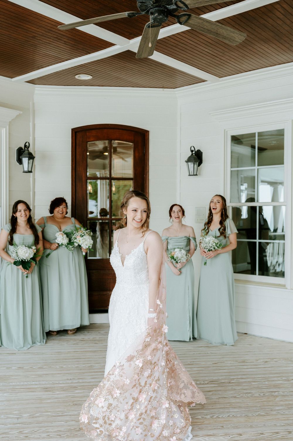 Bride in floral lace gown turns to reveal her dress as bridesmaids react with joy behind her.