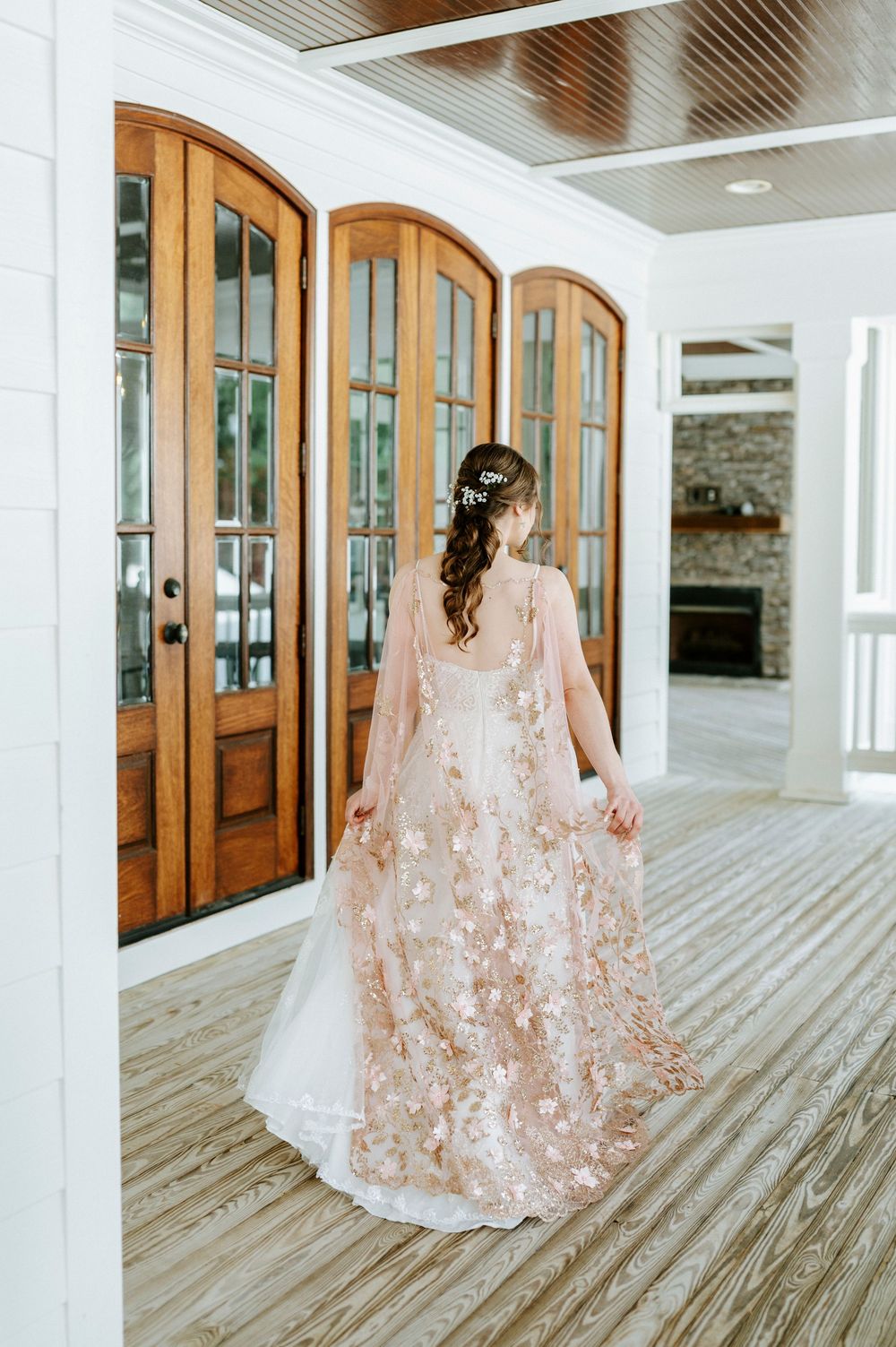 Bride in a stunning blush floral embroidered wedding gown with cape stands before arched wooden French doors.