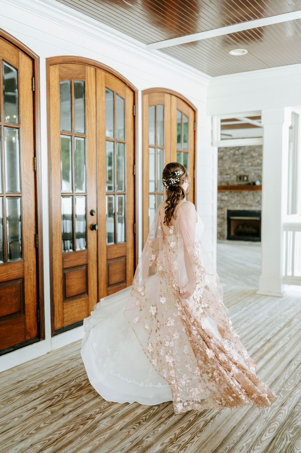 Bride twirling in a beautiful blush floral lace cape wedding dress on a porch with arched wooden doors.