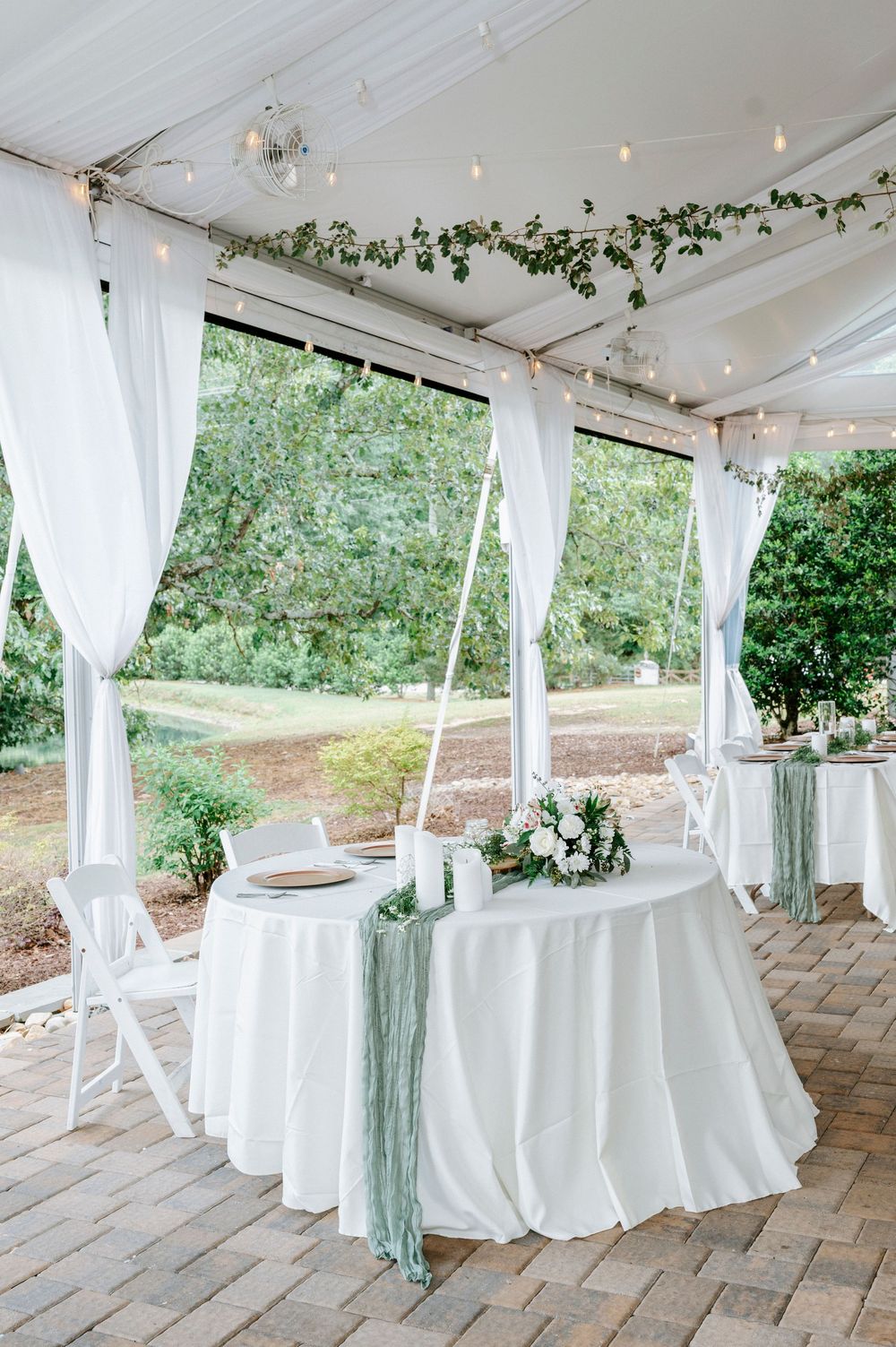 Elegant outdoor tented reception tables with white linens, sage runners, and string lights.