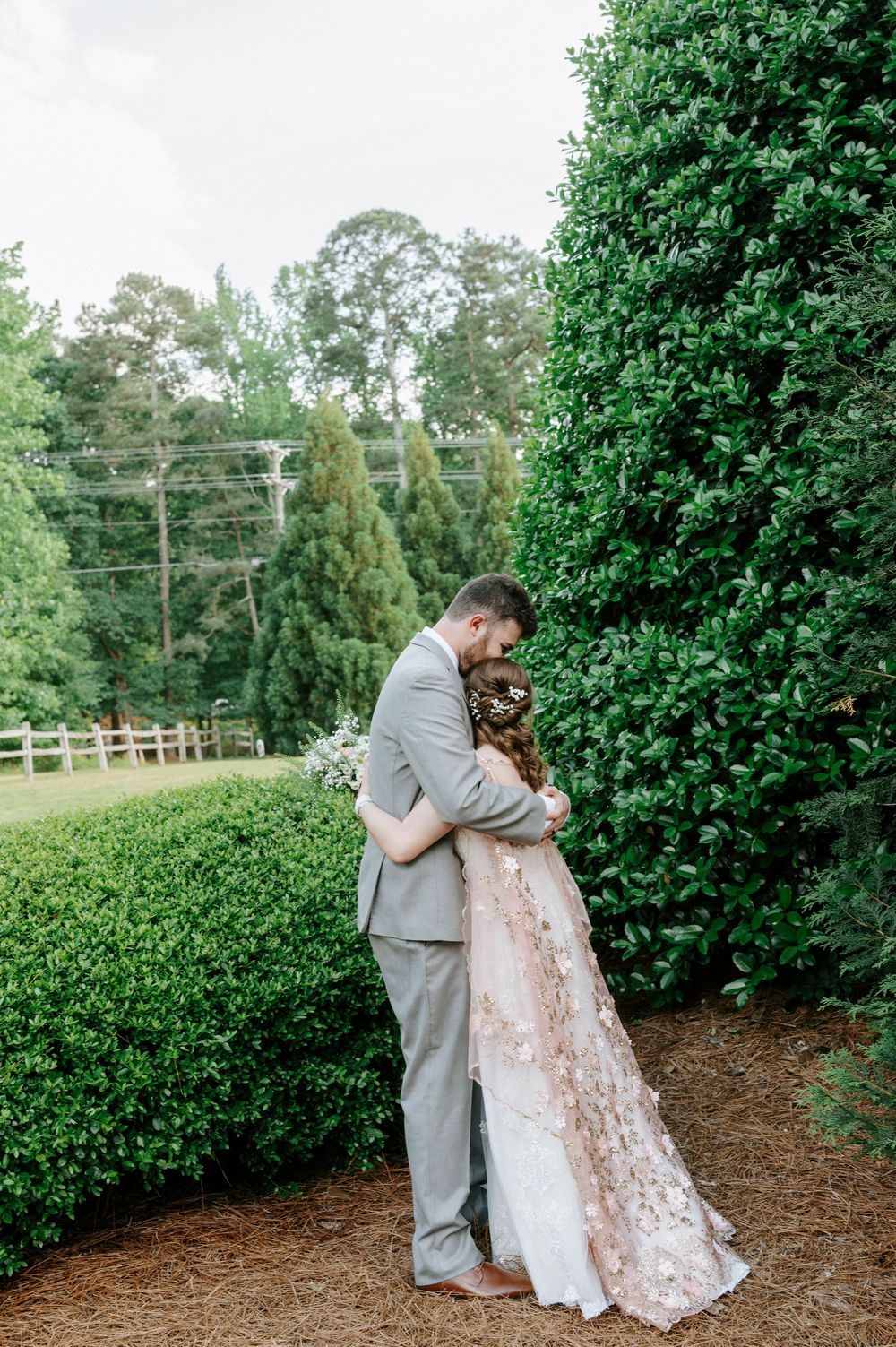 Bride and groom share a romantic embrace near lush green hedges during an outdoor wedding photo session.