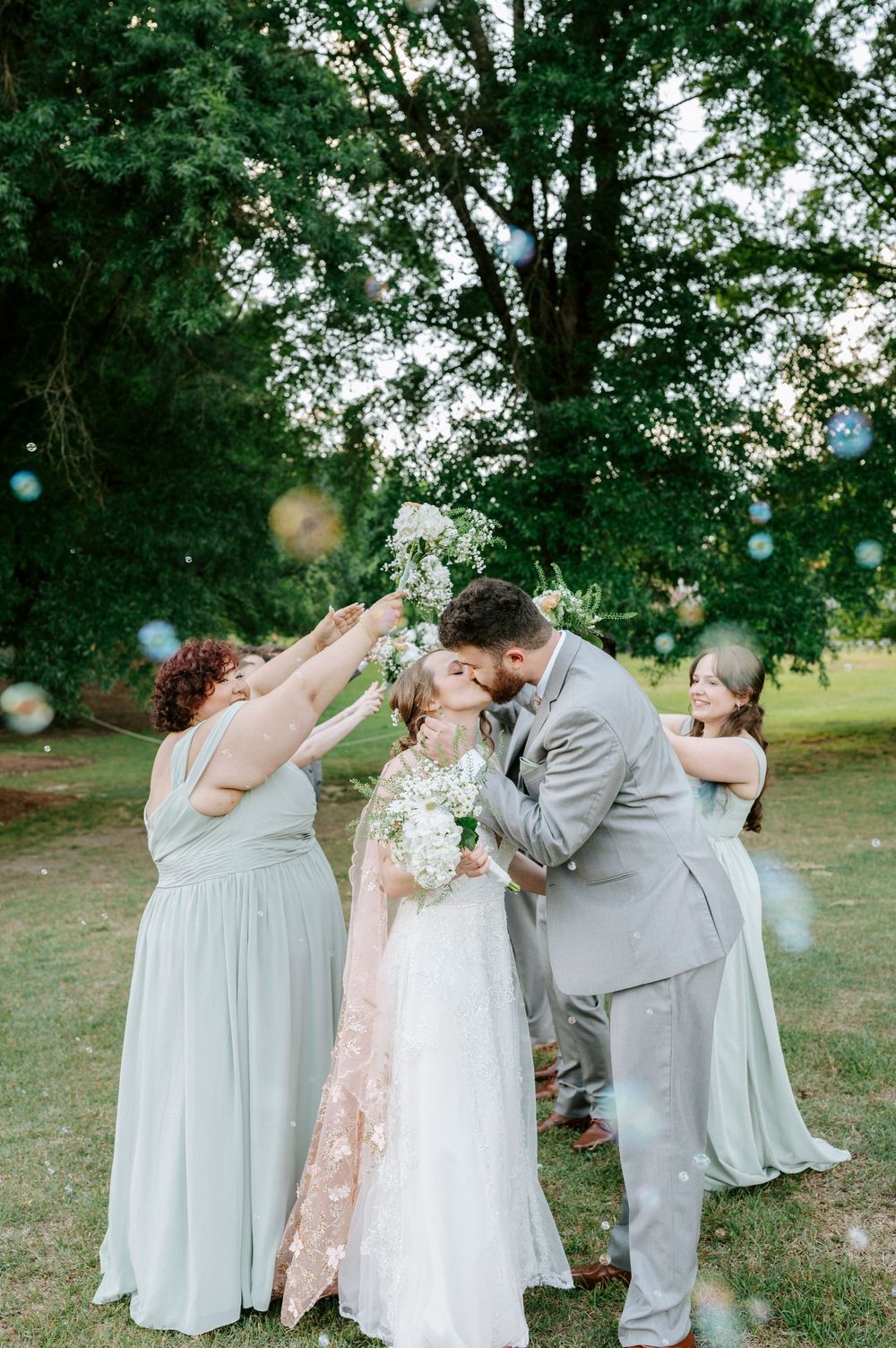 Bride and groom kissing surrounded by bridesmaids in sage green dresses with bubbles floating at outdoor wedding.