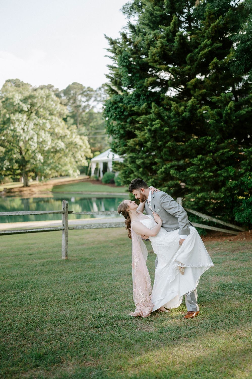 Groom dips bride for a romantic kiss on a lush green lawn with a pond and gazebo in the background.