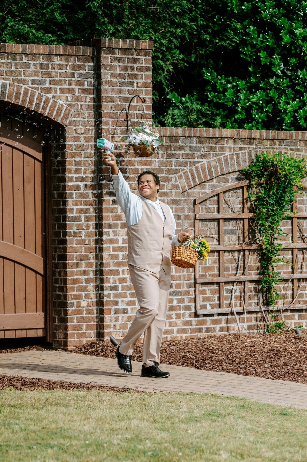 Cheerful flower girl attendant in beige suit carrying a basket of flowers at a brick garden entrance.