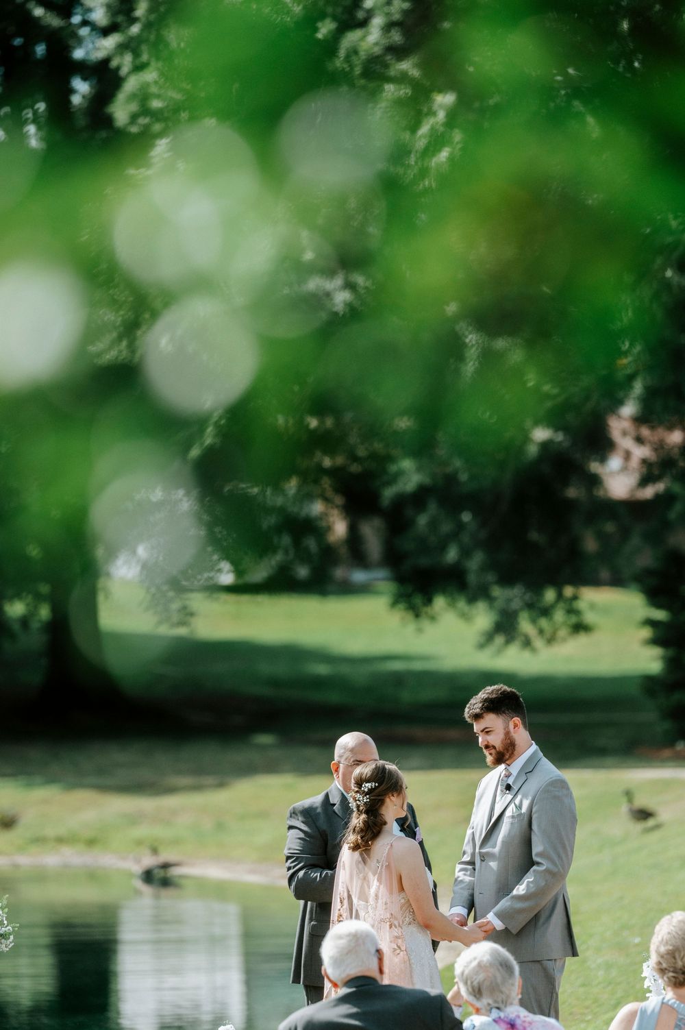 Groom in gray suit holds bride's hands during outdoor wedding ceremony by a pond surrounded by lush green trees.