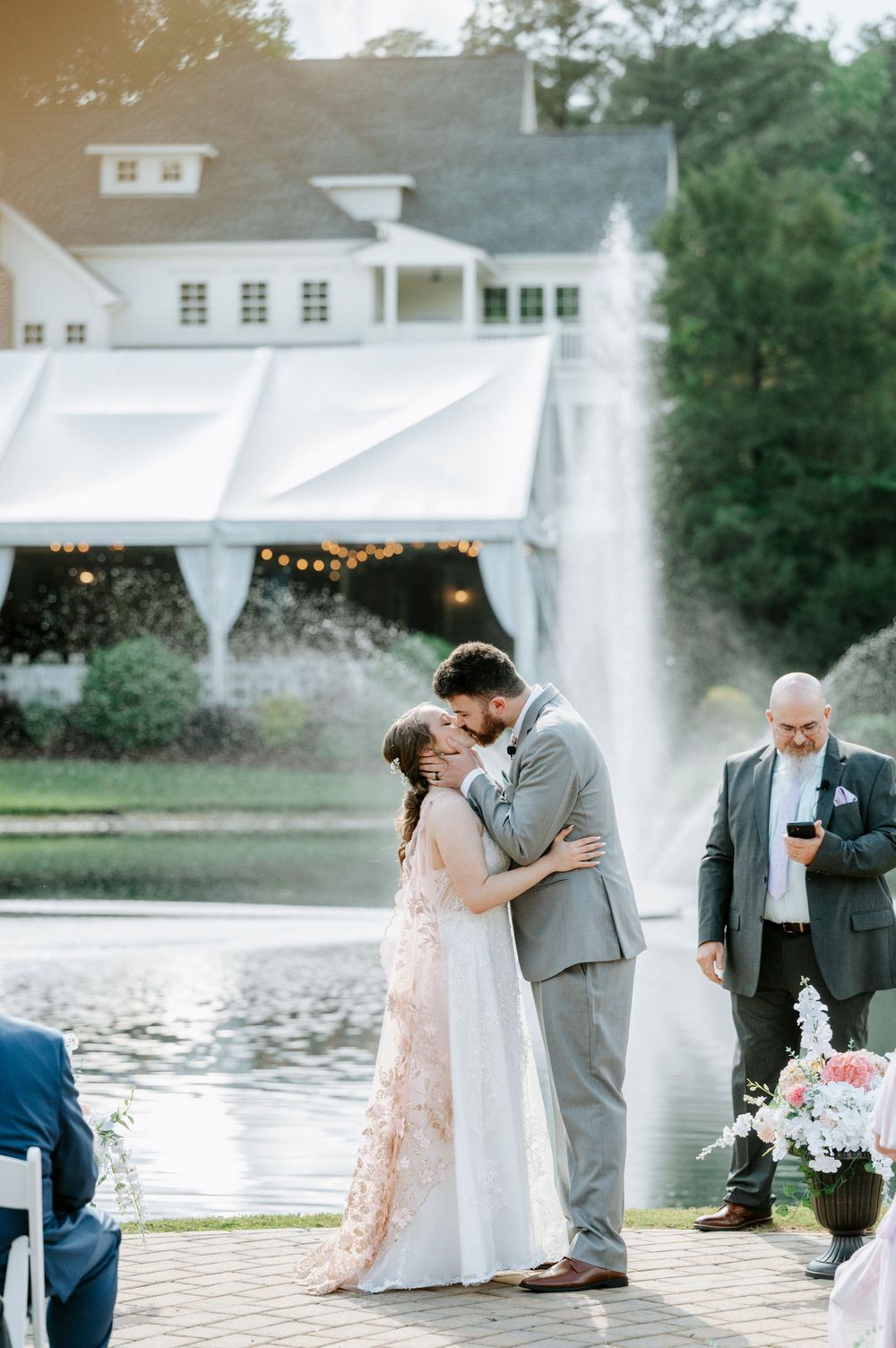 Bride and groom share first kiss at outdoor wedding ceremony beside a fountain with white venue in background.