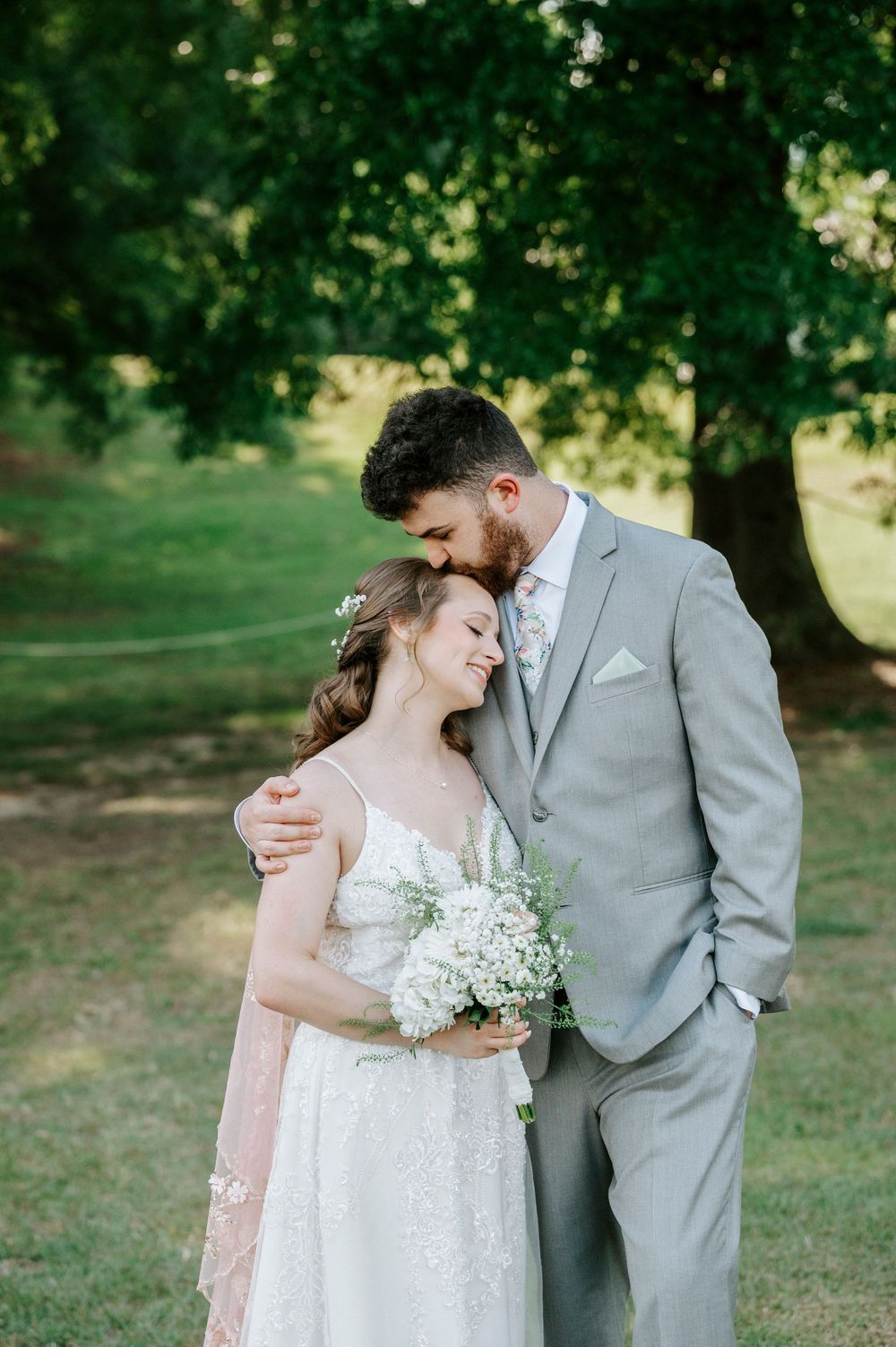 Groom in gray suit kisses bride on forehead as she holds a white bouquet under a large oak tree.