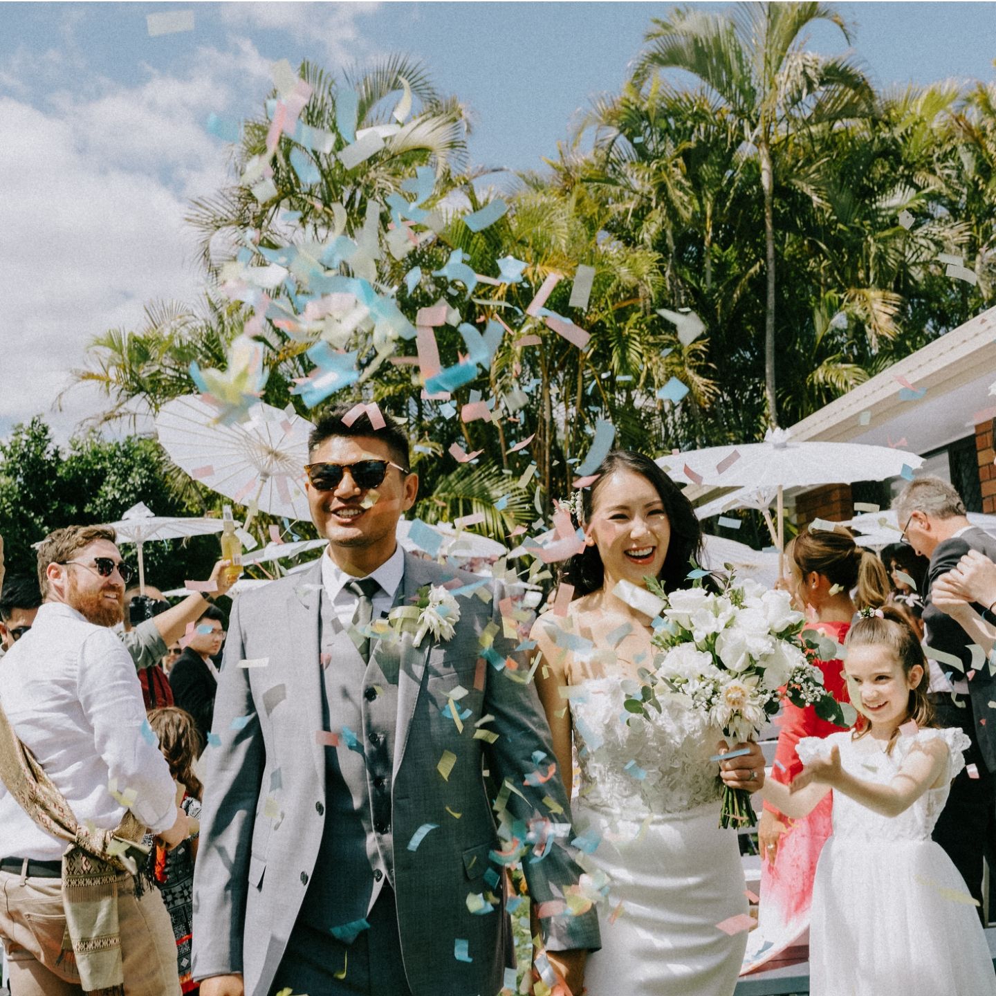 A joyful couple walks through colorful confetti at their outdoor wedding, surrounded by guests and a flower girl.