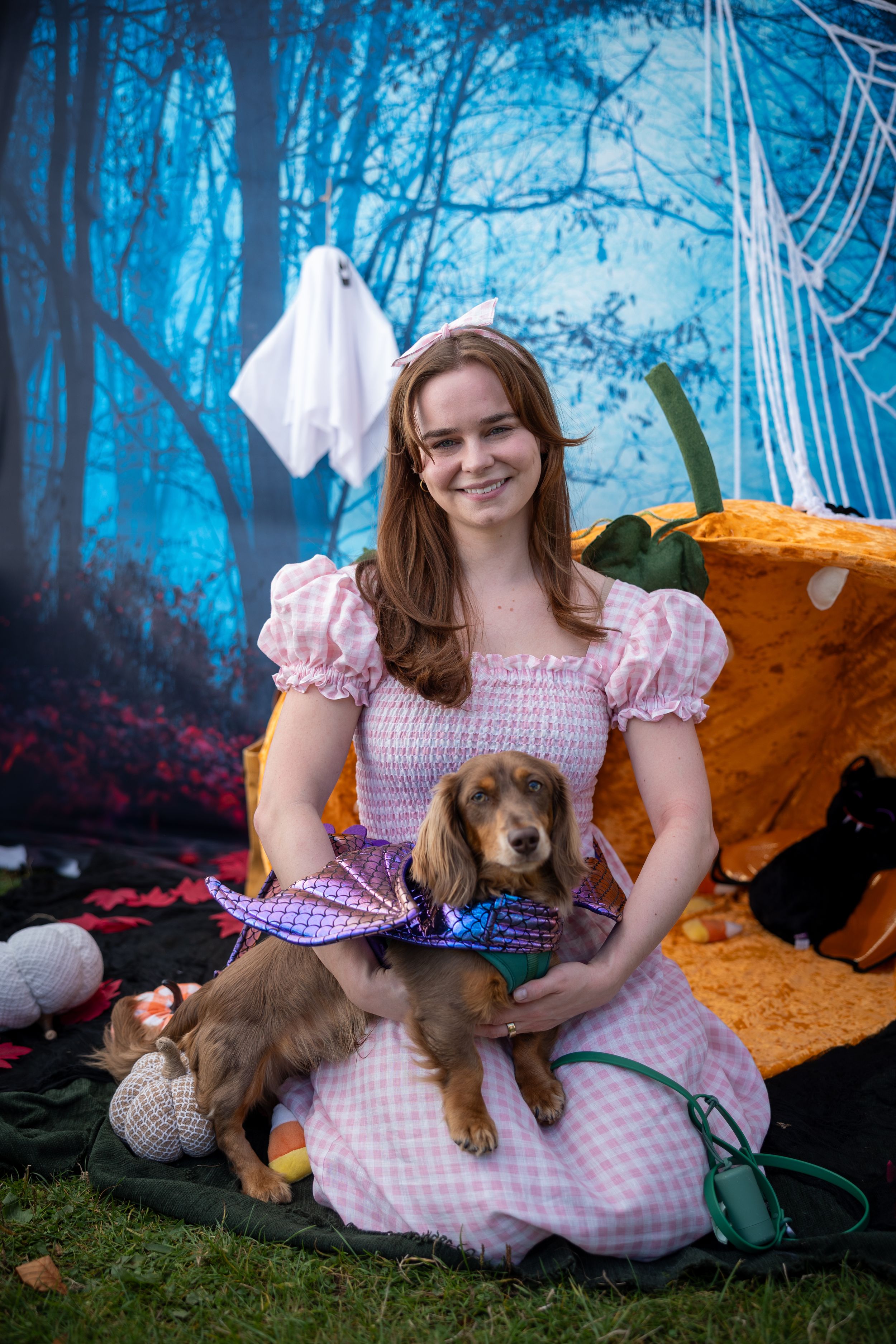 Dog dressed as a dragon sits with owner in a princess dress against spooky Halloween backdrop with pumpkins and ghosts.
