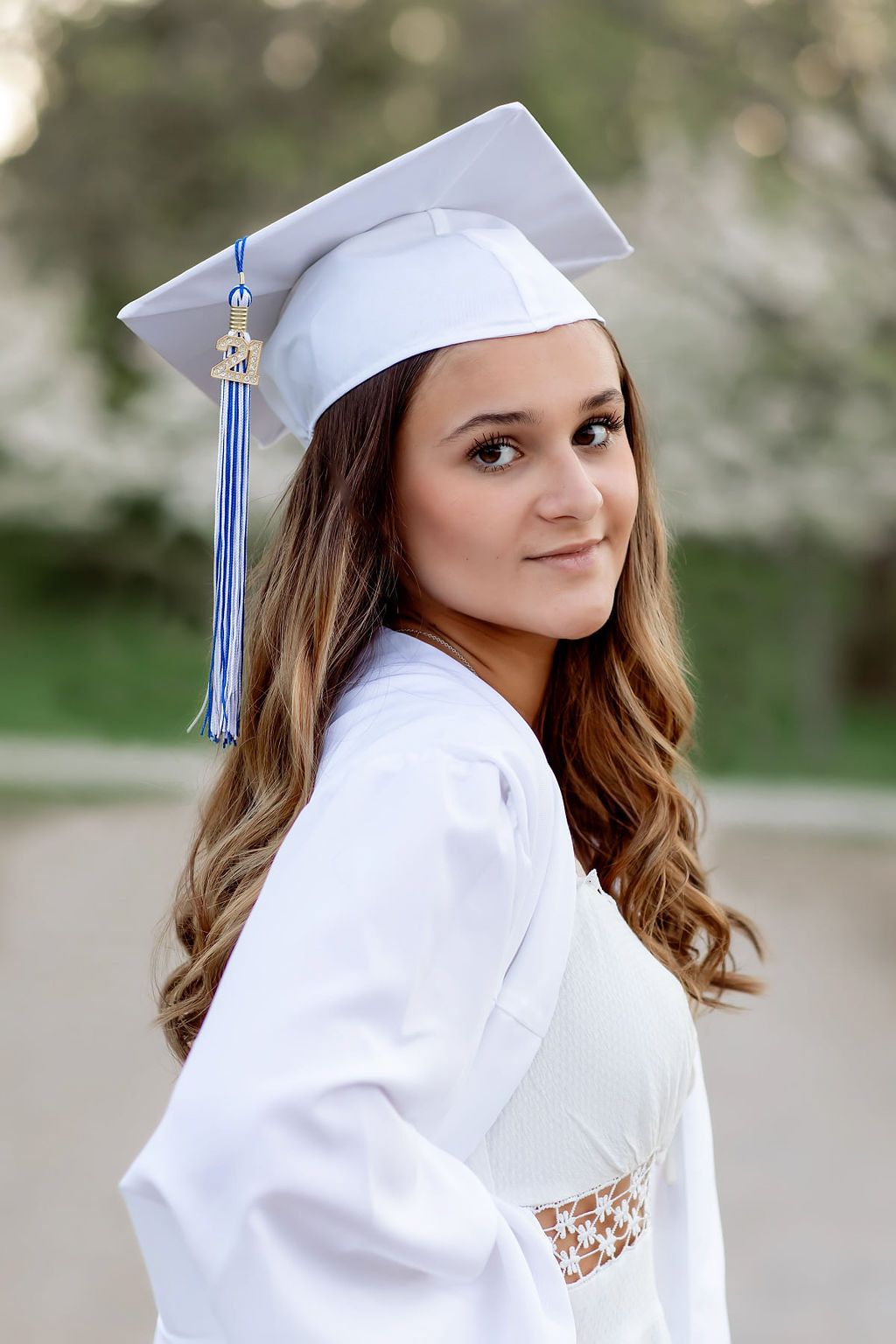 Graduate posing outdoors in white cap and gown with blue tassel against blurred natural background.