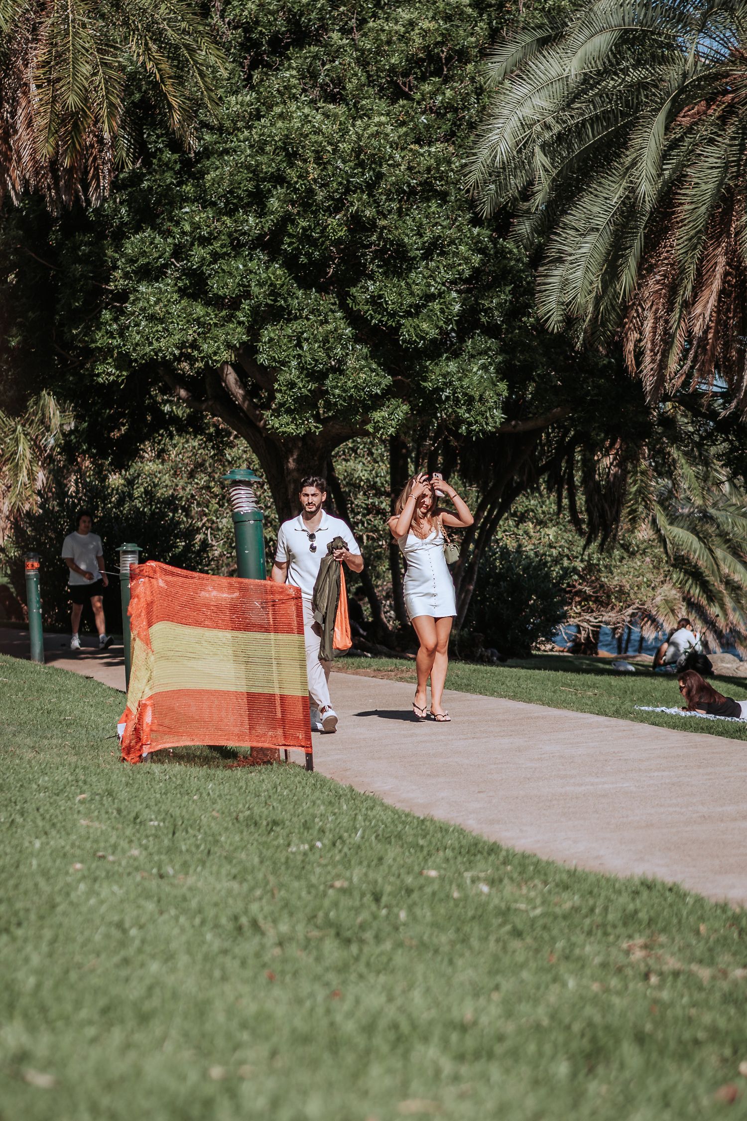 Couple walking along a park path on a sunny day with palm trees and a red-orange blanket.