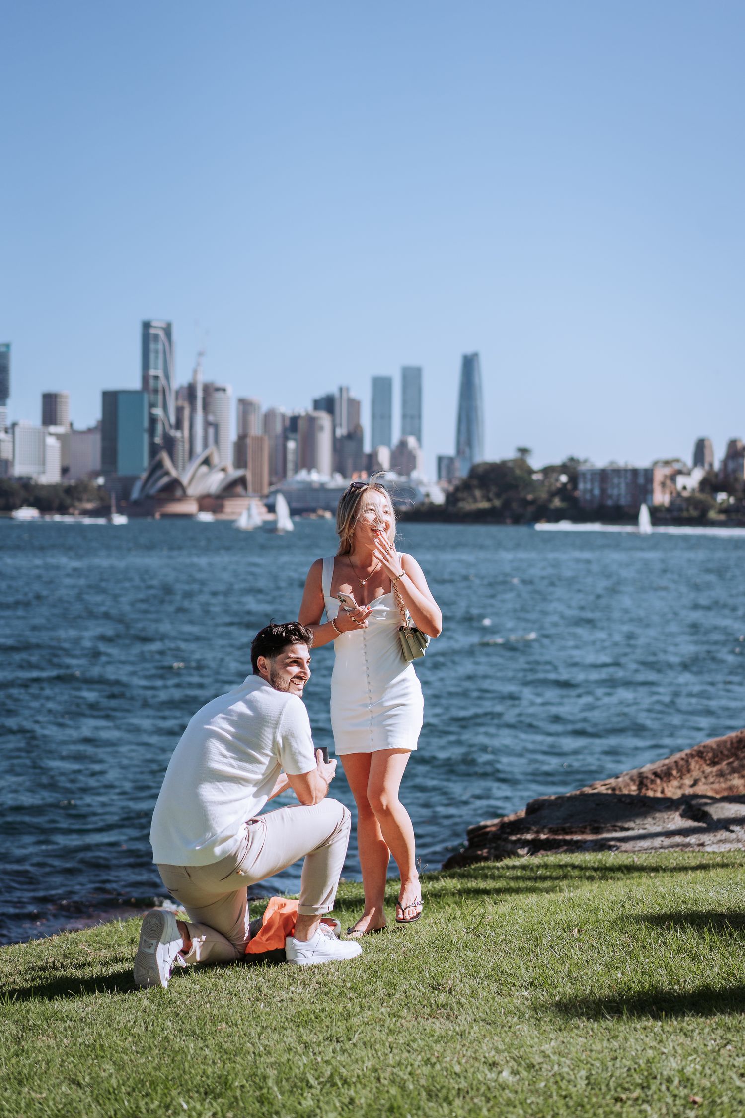 A romantic proposal taking place on a grassy waterfront with the Manhattan skyline in the background on a sunny day.