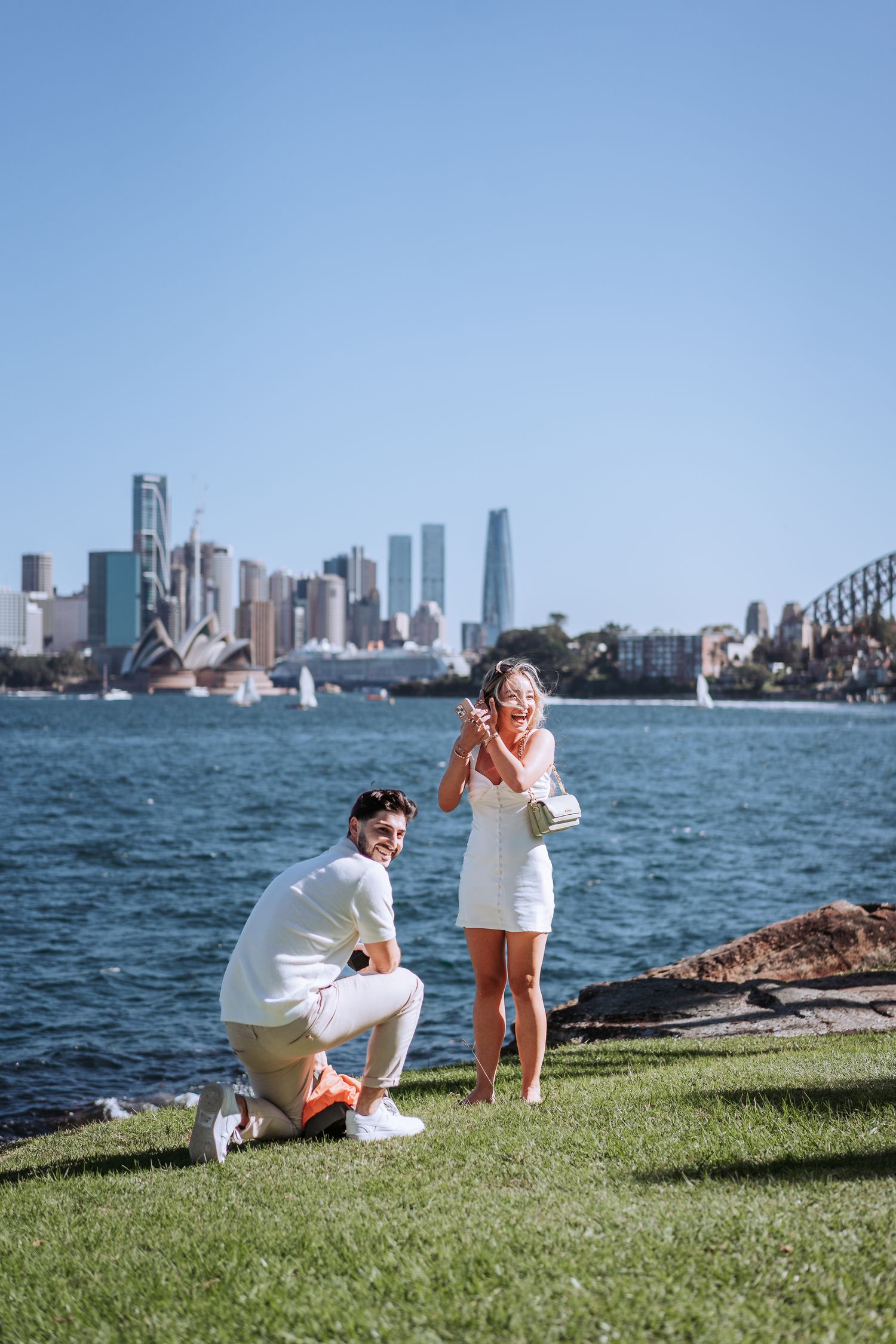 A couple shares a touching moment against the iconic Sydney Opera House and harbor skyline on a sunny day.