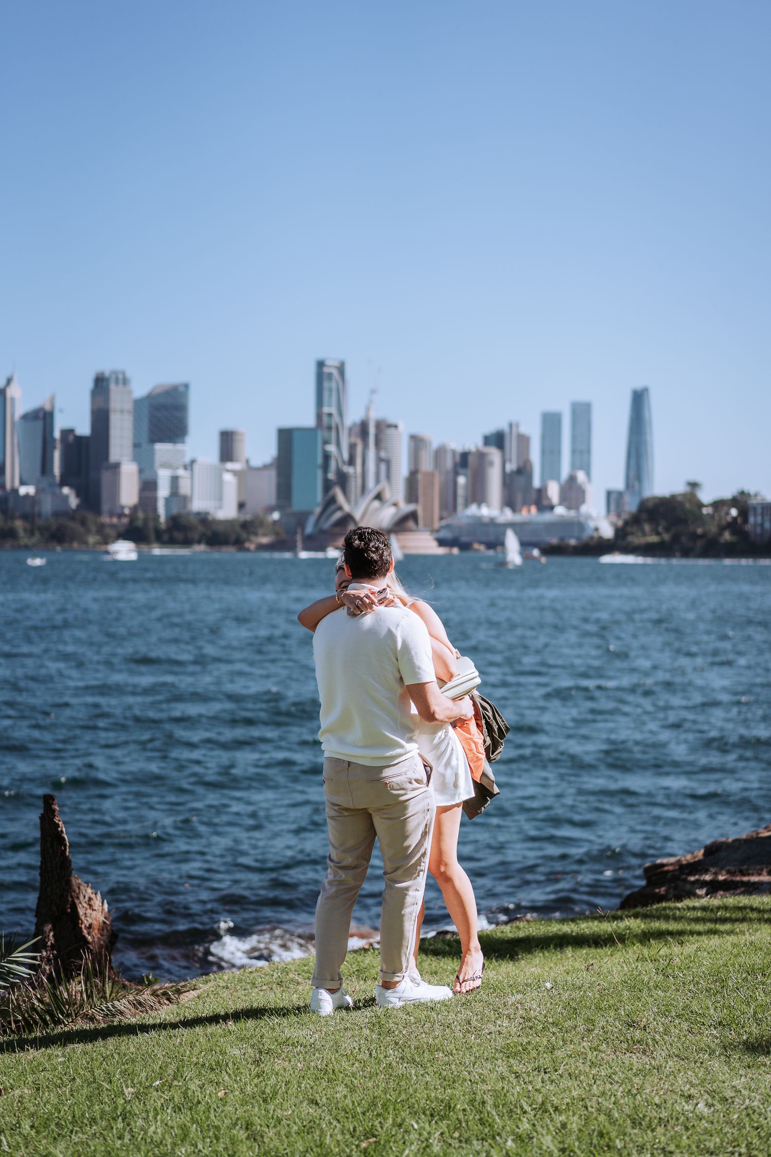 A couple embraces while enjoying views of Sydney's skyline and harbor from a grassy viewpoint on a sunny day.