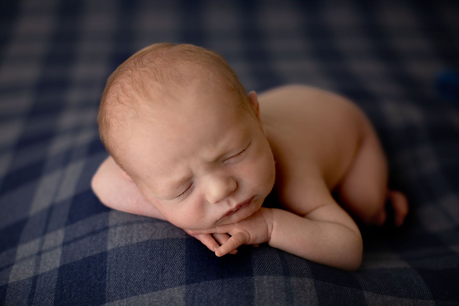 Sleeping newborn baby curled up on dark blue plaid blanket in peaceful newborn photography pose.