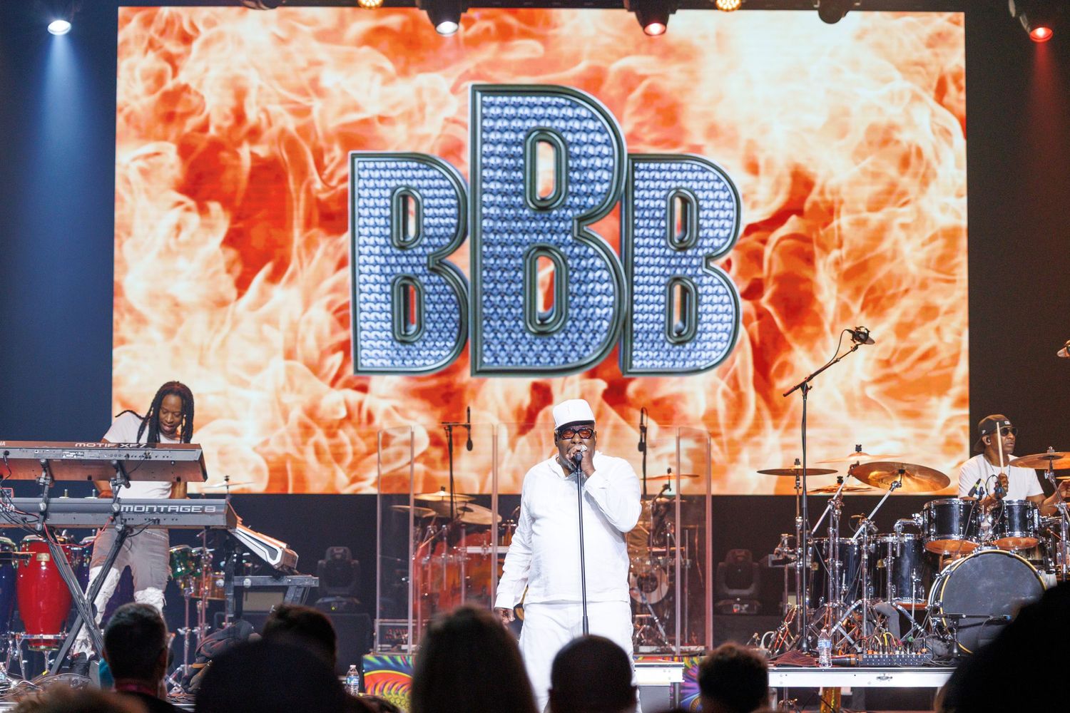 A performer in white attire stands on stage in front of large illuminated BBB letters with orange flames in the background.