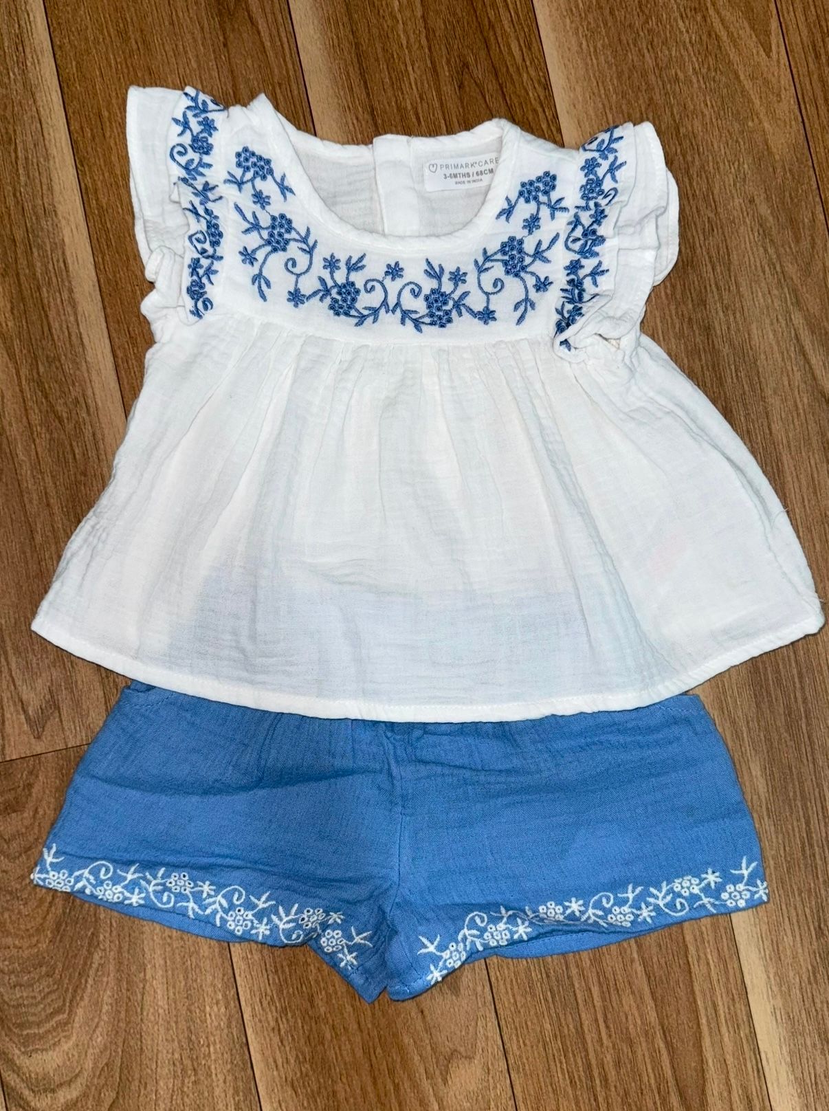White cotton baby top with blue embroidery paired with denim shorts shown from various angles.