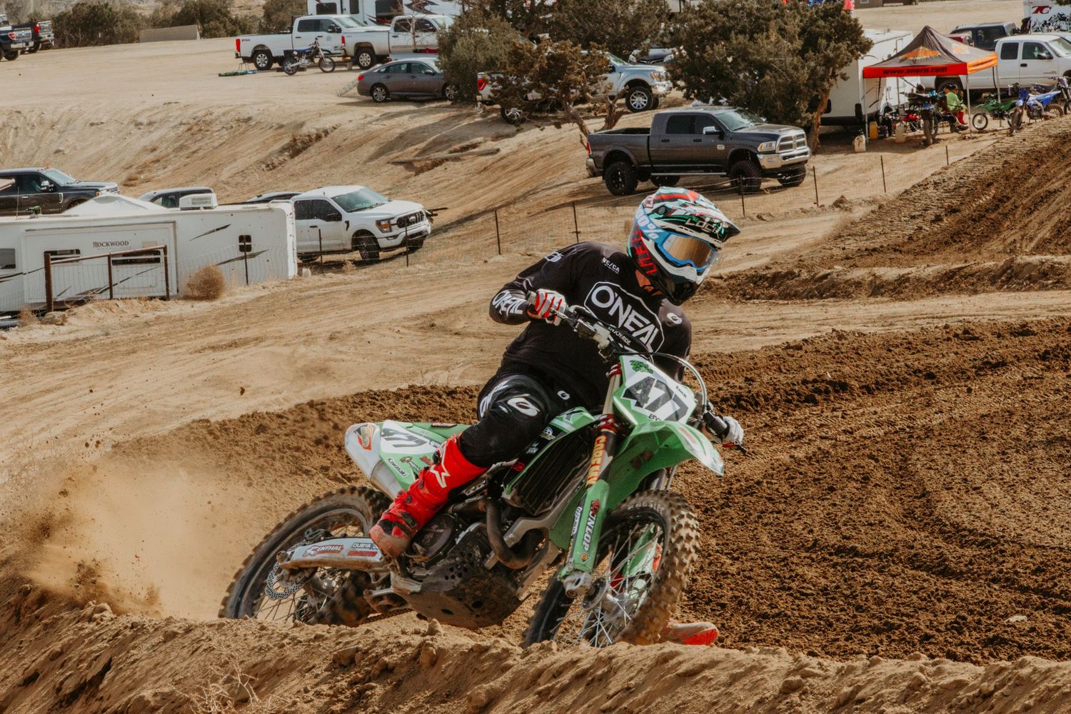 A dirt bike rider navigates a steep motocross track turn while spectators and parked cars watch from above.
