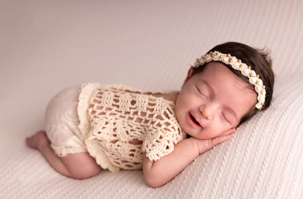 A sleeping newborn in a cream-colored lace wrap poses on a soft pink backdrop.