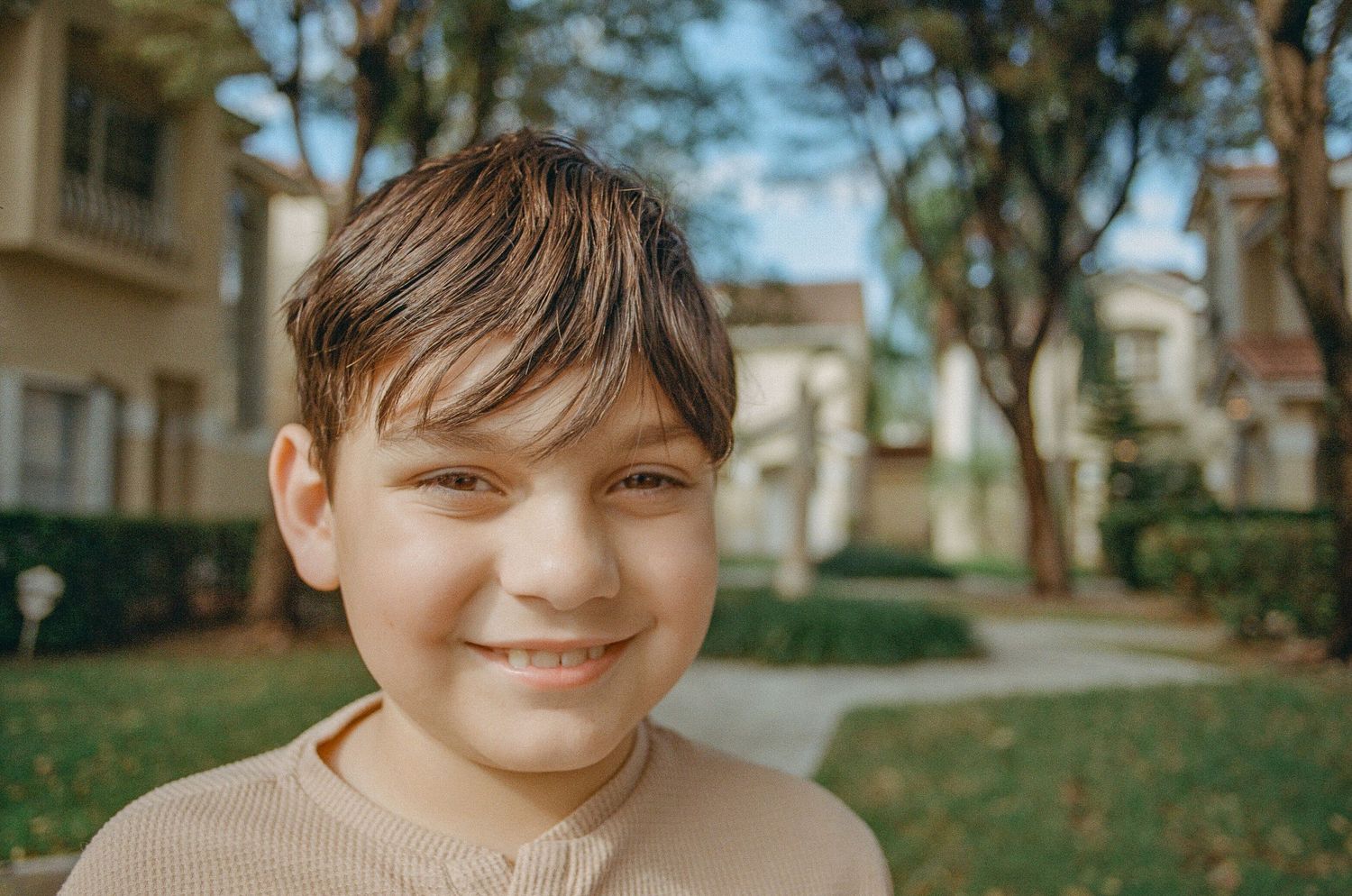 A young child in a beige sweater smiles warmly while standing outdoors near residential buildings on a sunny day.